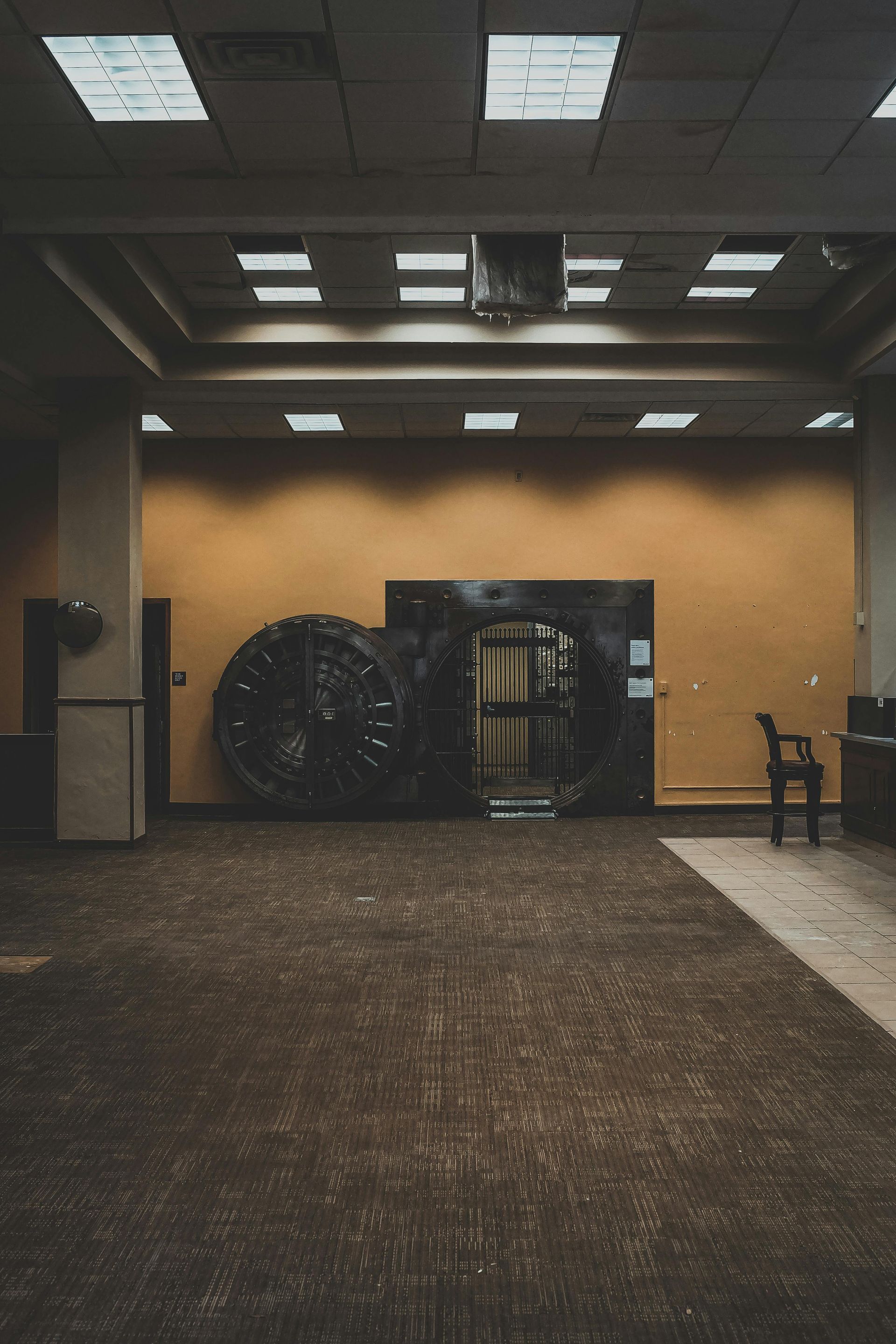 A heavy circular steel bank vault door stands open in an otherwise empty, dimly lit room with tan walls and carpet.