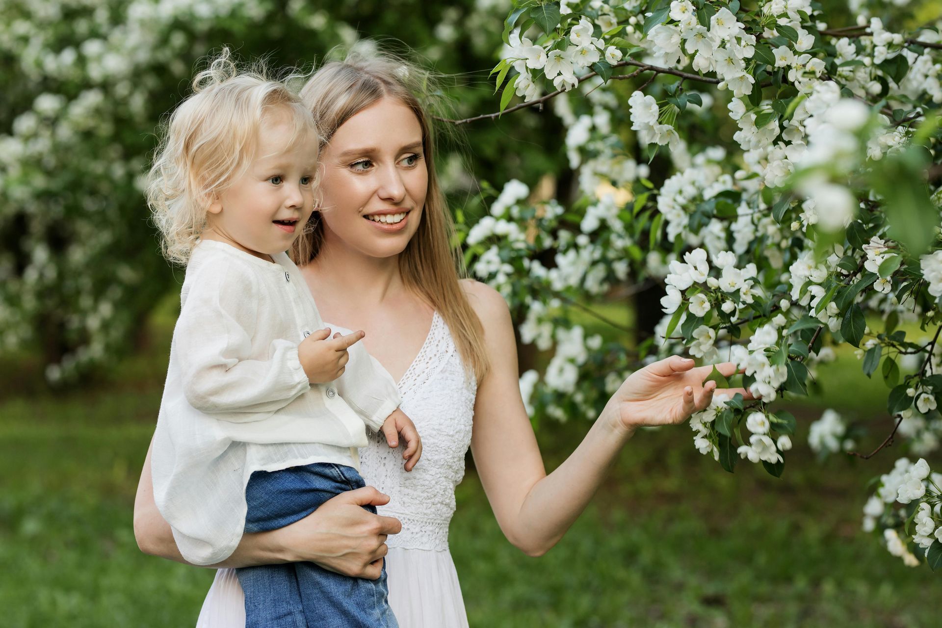 A smiling person holding a child in a garden with blooming white trees.