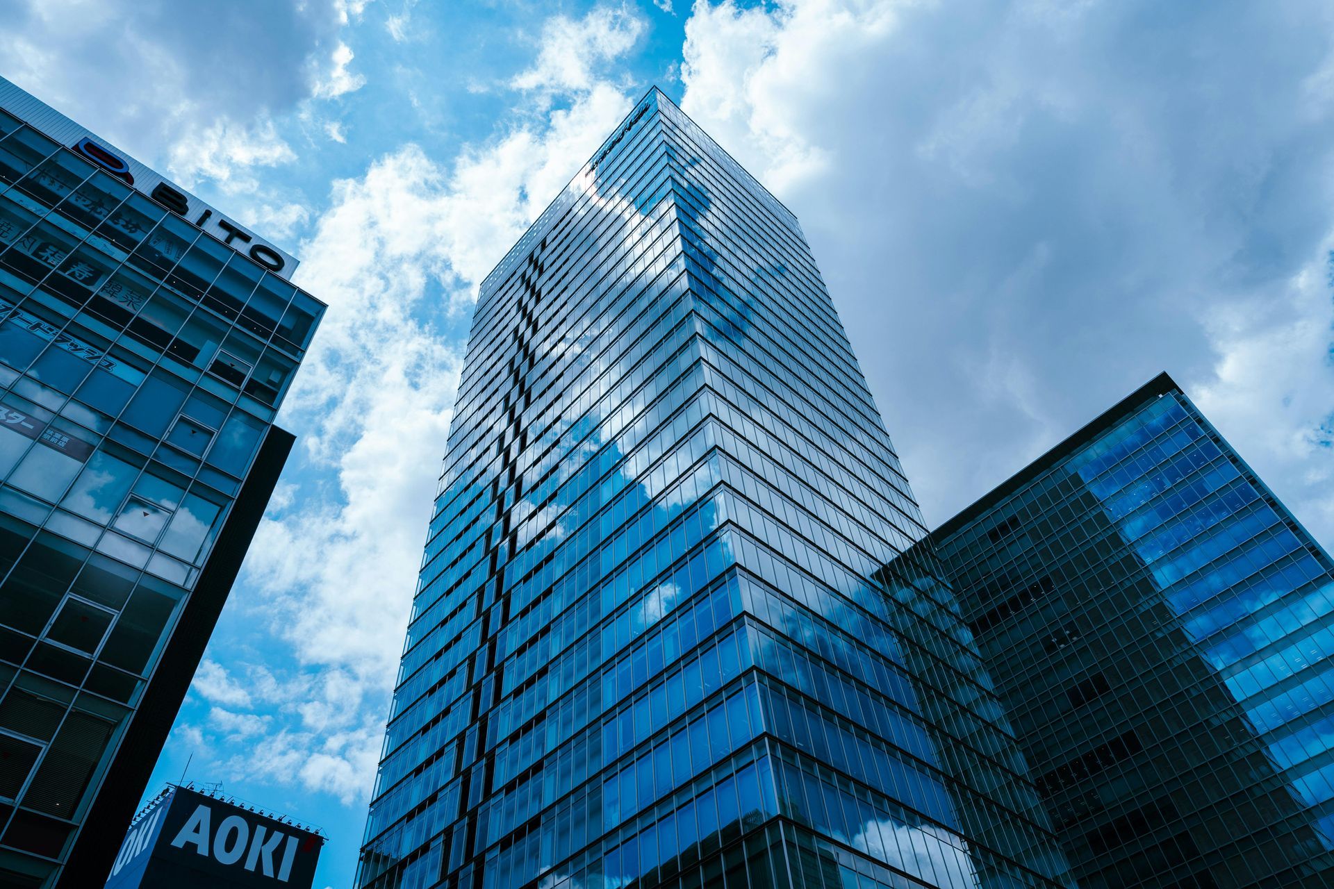 Tall glass skyscrapers reflect a blue, cloudy sky from a low-angle perspective, with an 
