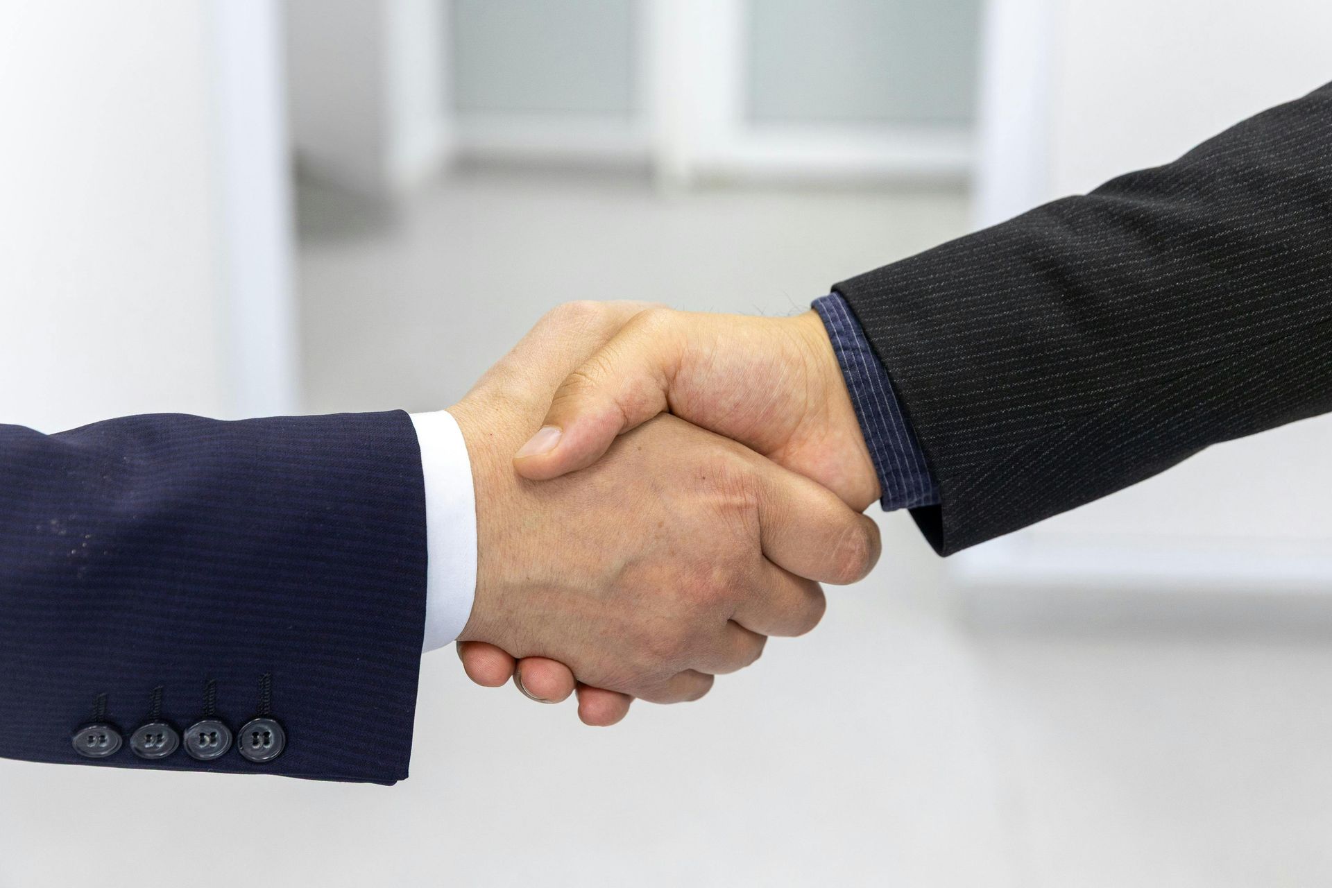 Two business professionals in dark suit jackets shaking hands against a bright, blurred office background.