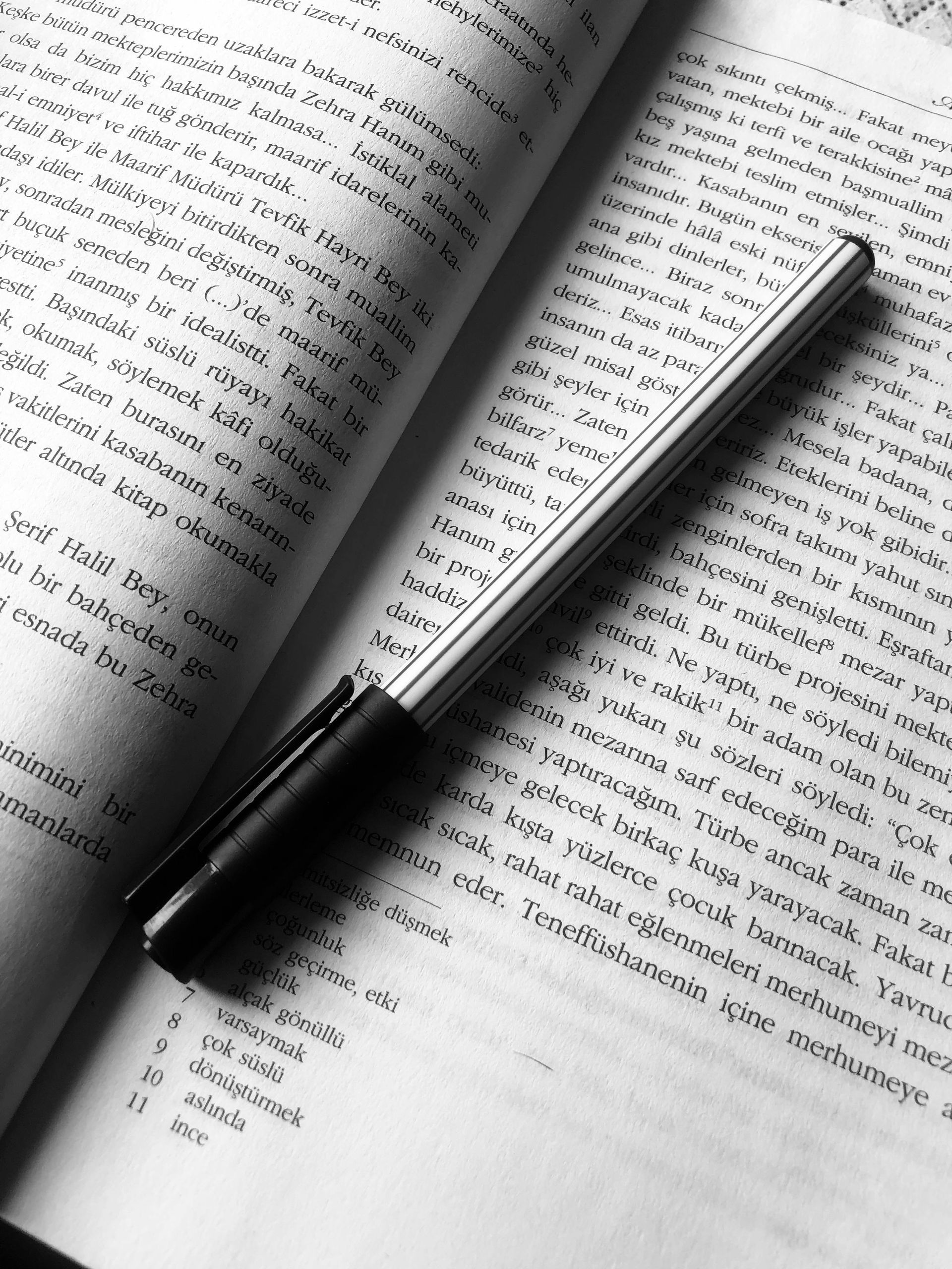 A pen rests diagonally across an open book with printed text and a numbered list at the bottom.