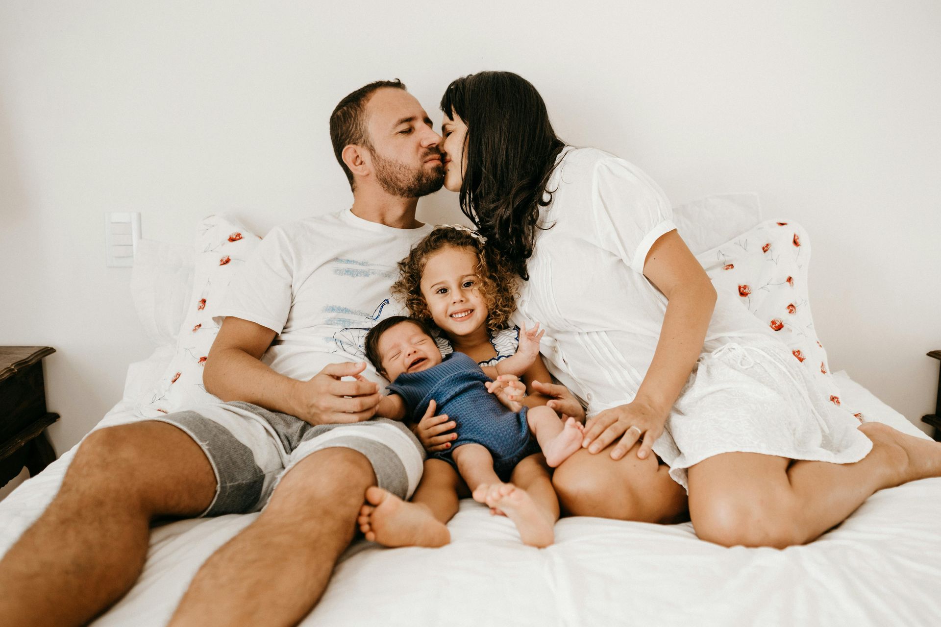 A family of four sits on a bed, with parents kissing while holding an infant and a young child between them.