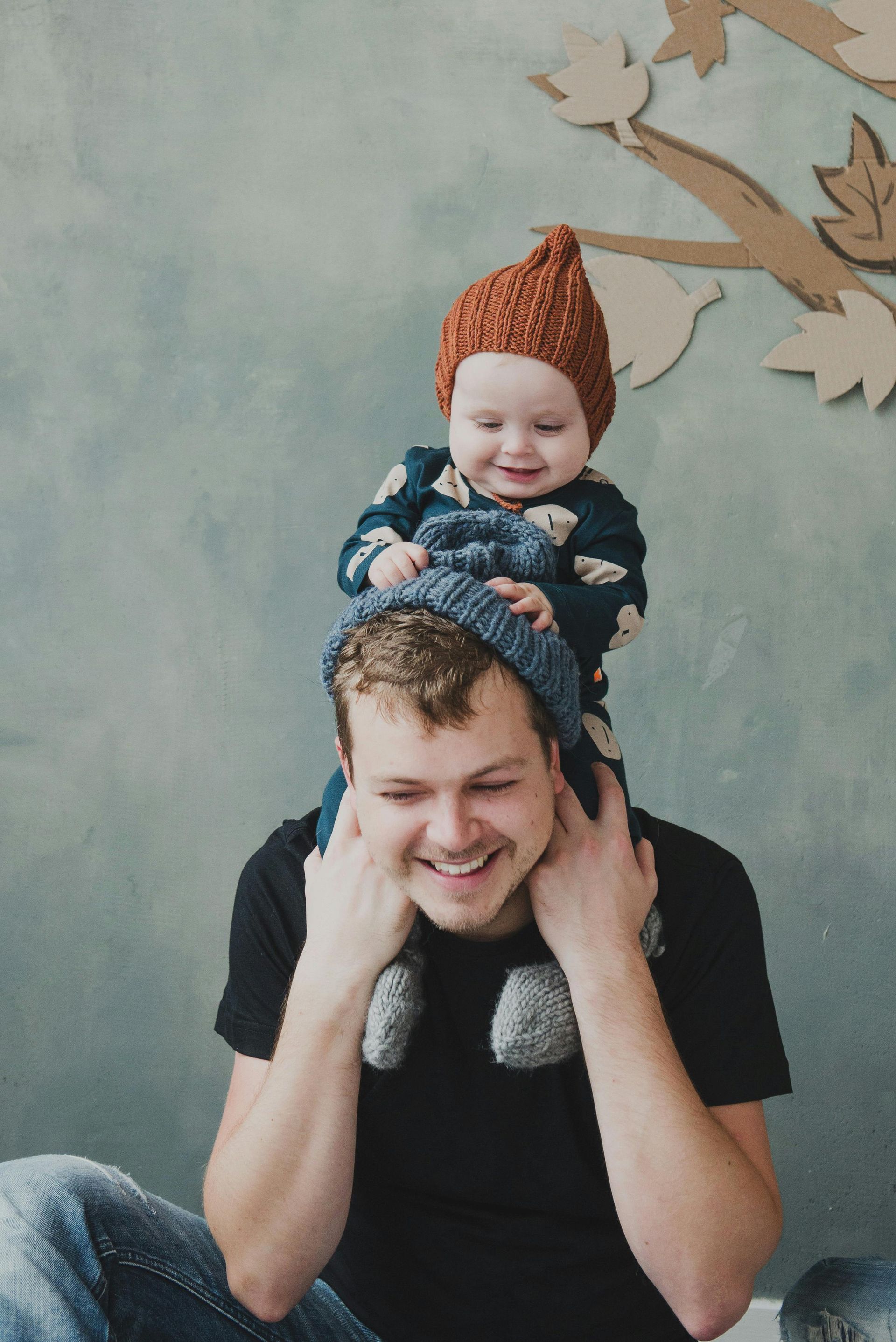 A man smiling while a baby sits on his shoulders, both wearing knit hats against a gray wall with a cardboard tree.
