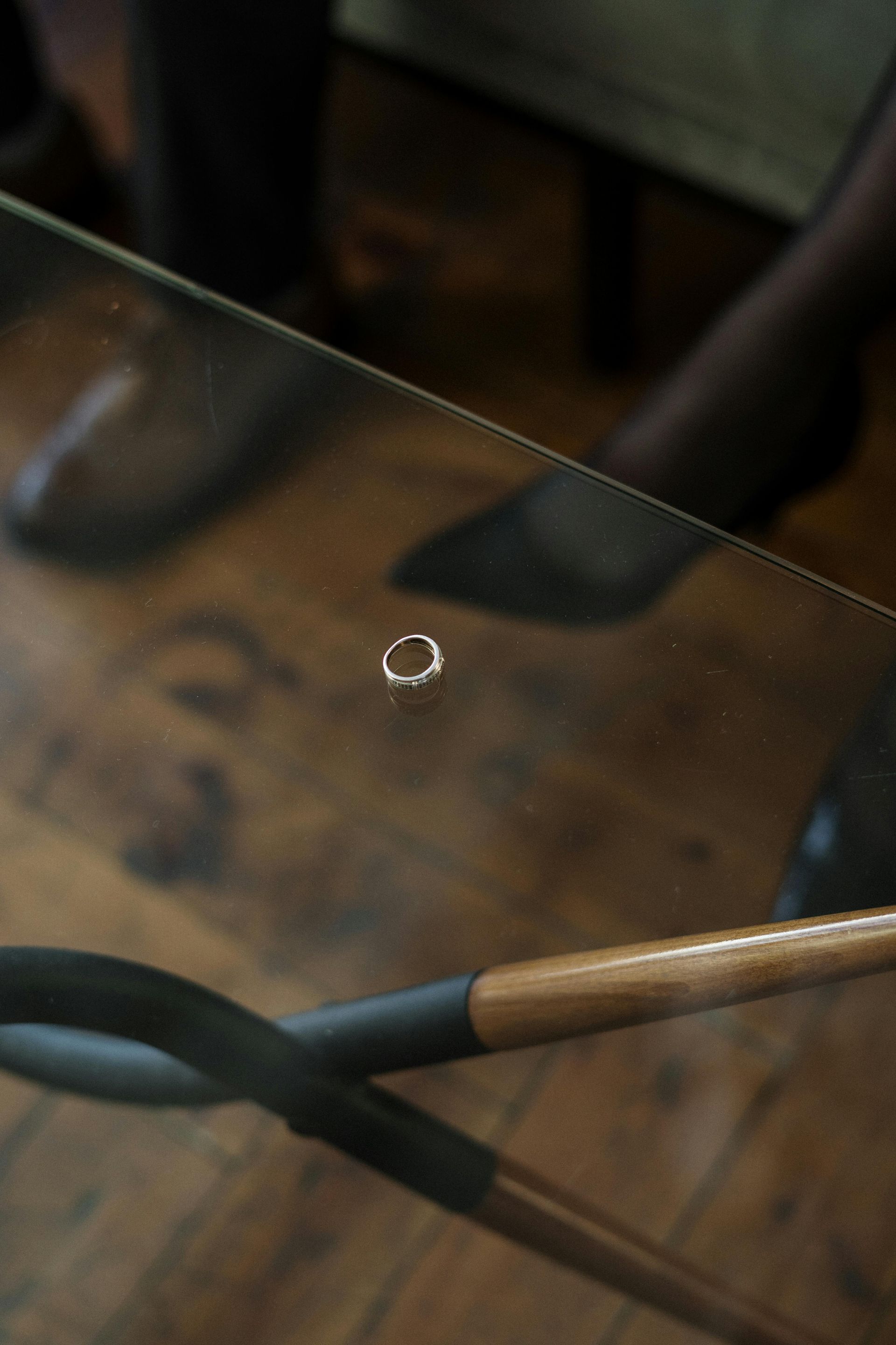 A small silver hoop earring lies on a glass table, with blurred feet and wooden furniture in the background.