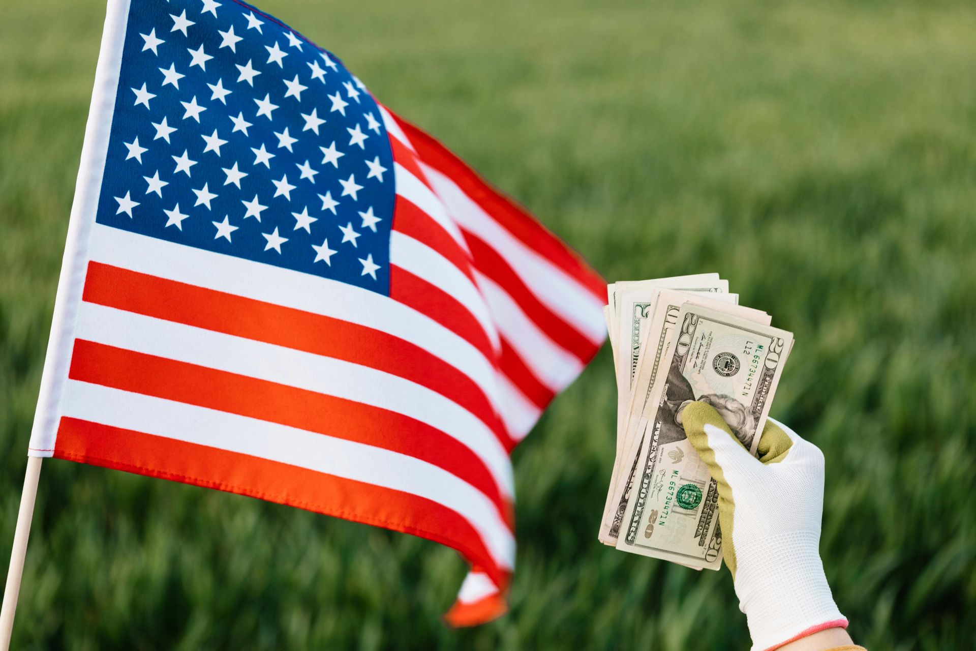 A hand wearing a white work glove holds a stack of U.S. dollar bills in front of an American flag in a grassy field.