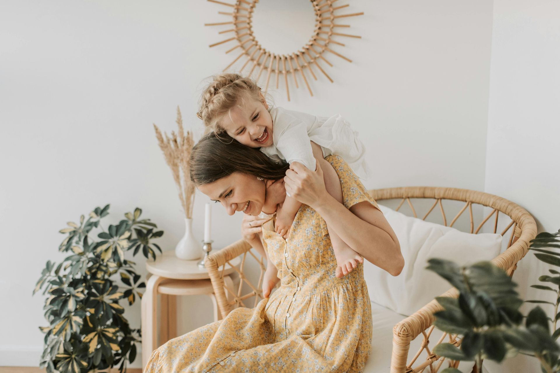 A person in a yellow dress sits in a rattan chair, holding a laughing child on their shoulders in a bright, cozy room.