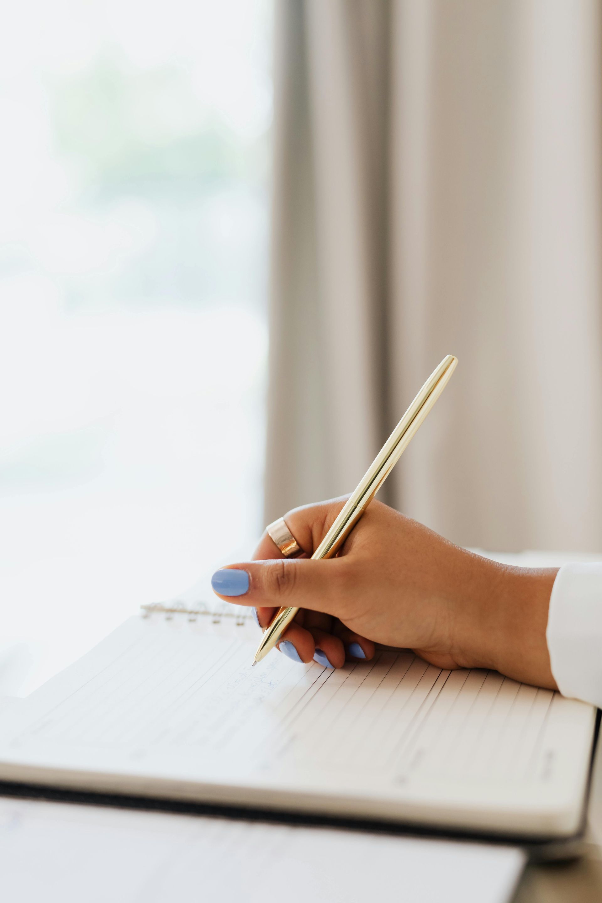 A hand with light blue nail polish writes in a notebook with a gold-textured pen against a soft, bright background.
