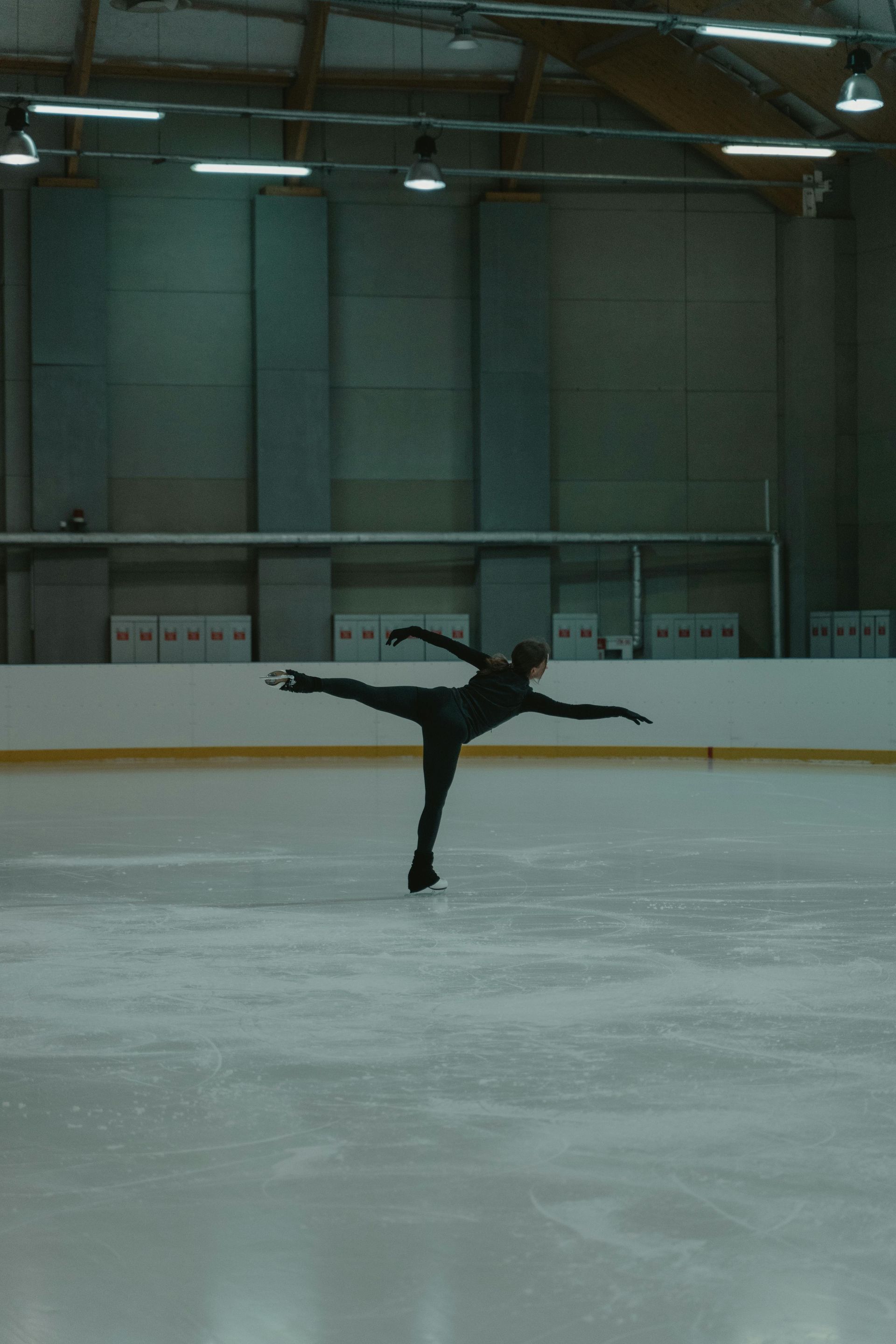 A person in black athletic gear performs an arabesque ice skating move in a brightly lit indoor ice rink.