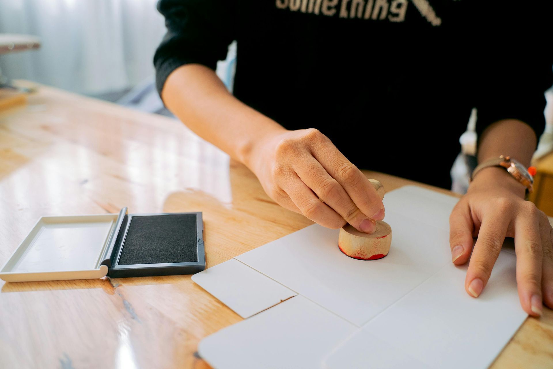 A person presses a round rubber stamp onto a sheet of white paper next to an open ink pad on a wooden table.