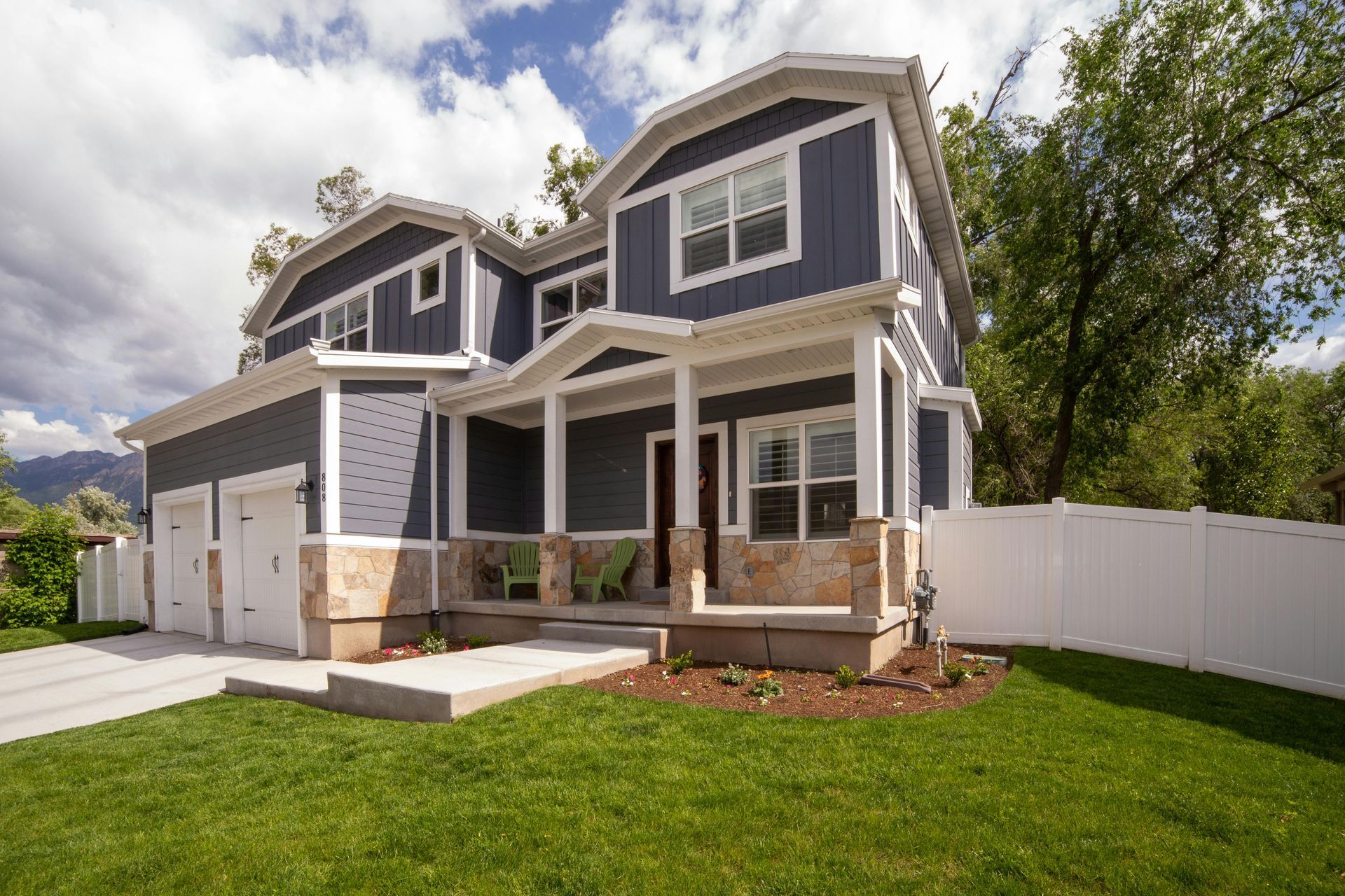 A two-story blue house with stone accents, a porch, a garage, and a white fence, surrounded by a lawn under a cloudy sky.