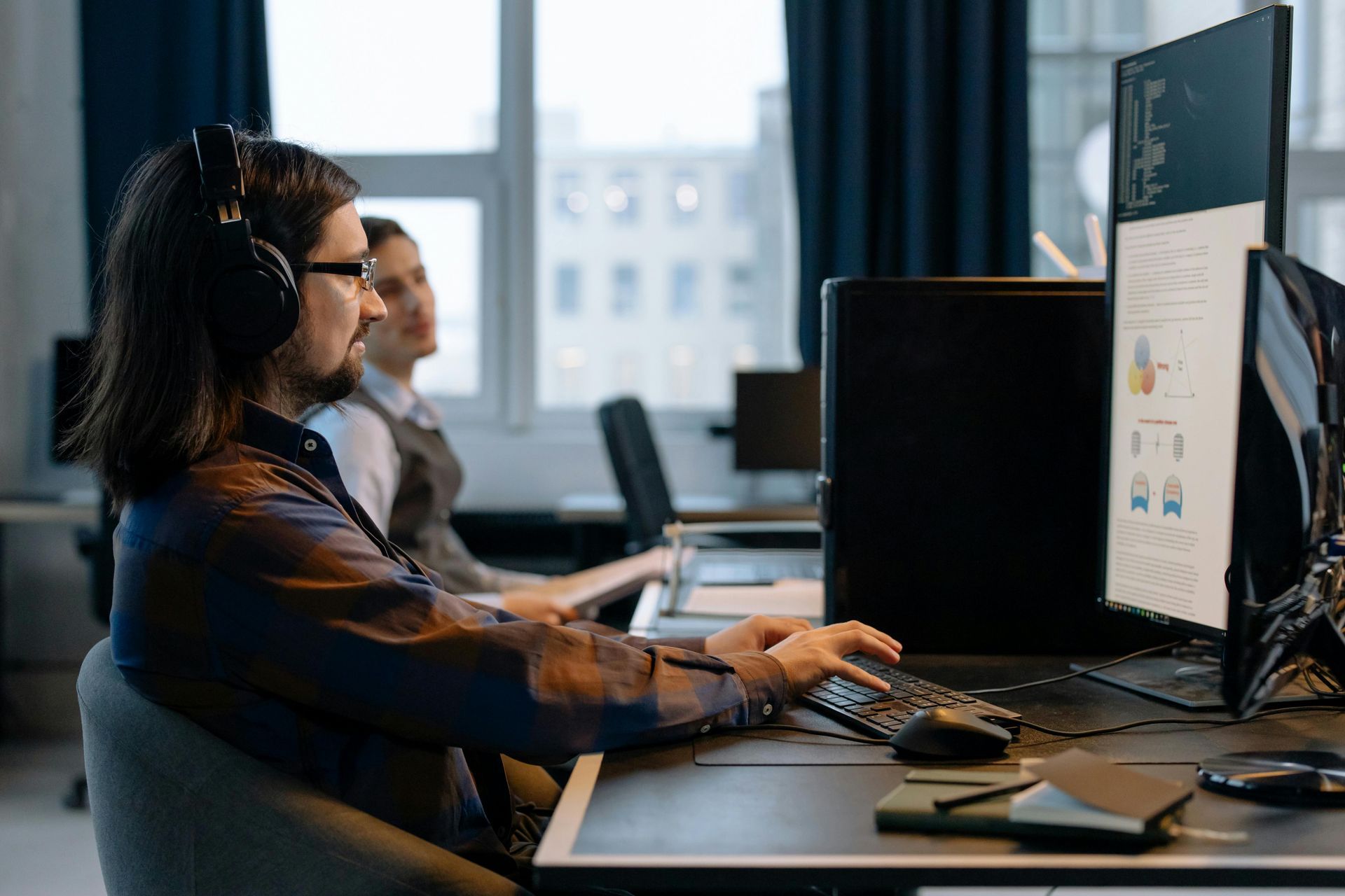 A person with headphones sits at a desk in an office, typing on a computer while another person sits in the background.