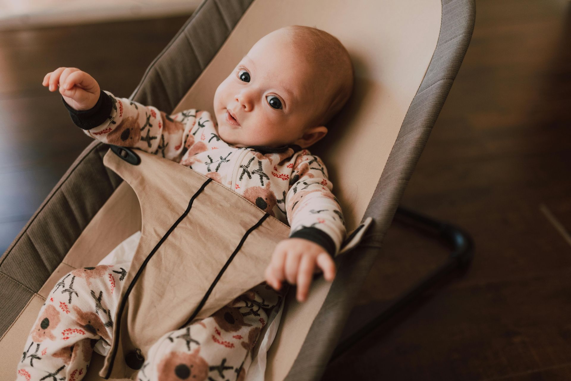 A baby in a patterned onesie sits in a beige-colored baby bouncer on a dark floor, looking toward the camera.