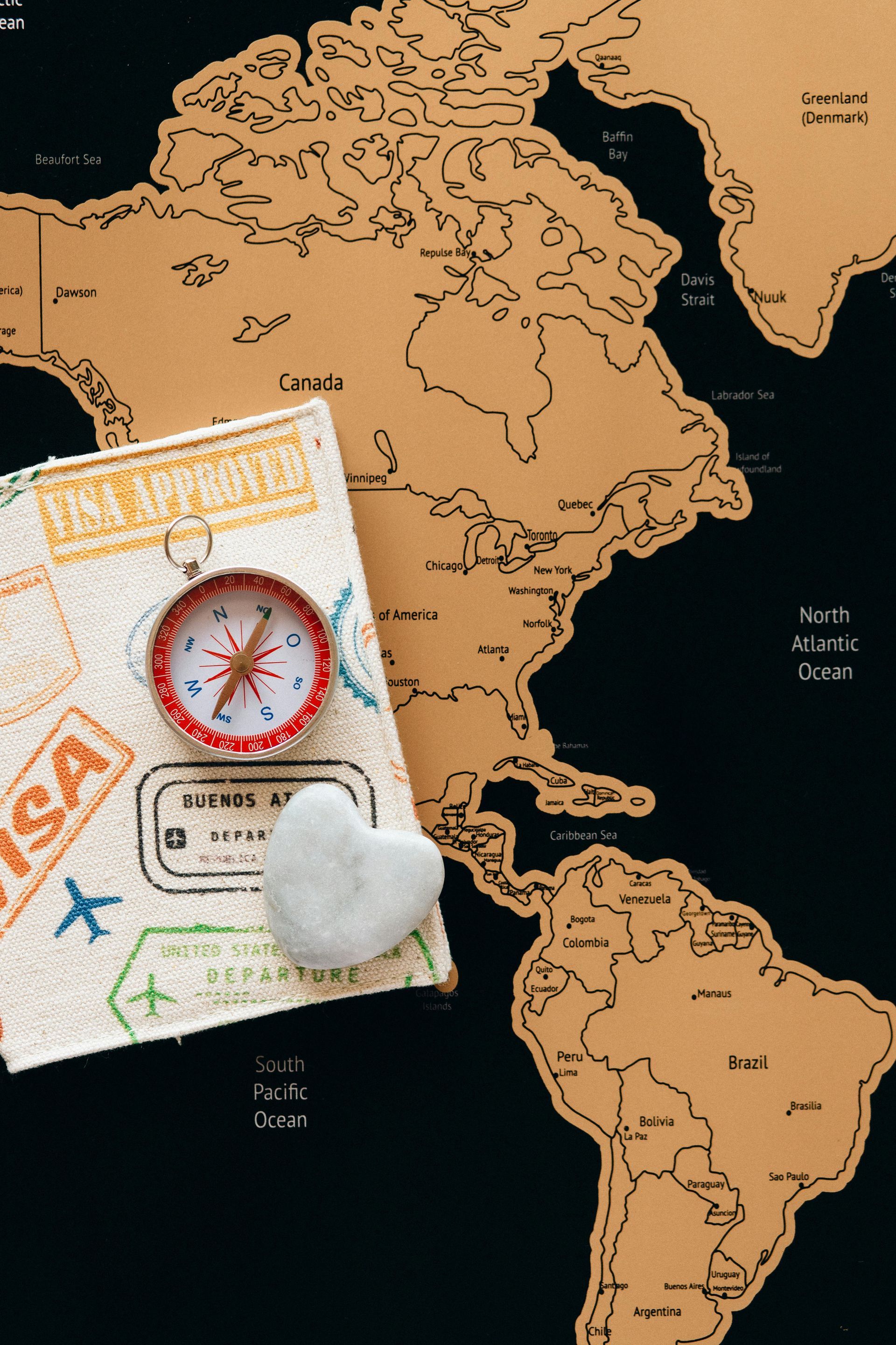 A vintage compass and a heart-shaped stone rest on a passport over a scratch-off map of the Americas.