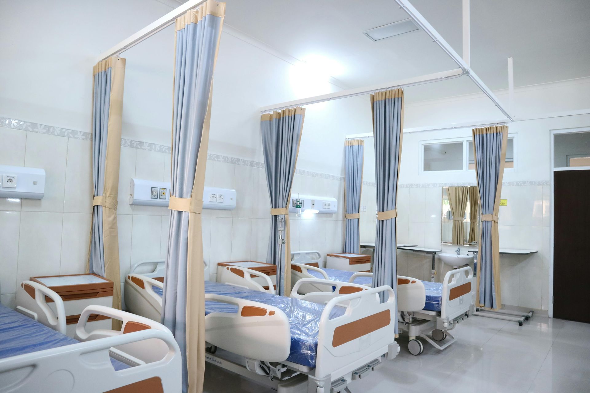 Hospital ward with empty medical beds, blue curtains, and wall-mounted equipment in a bright, clean room.