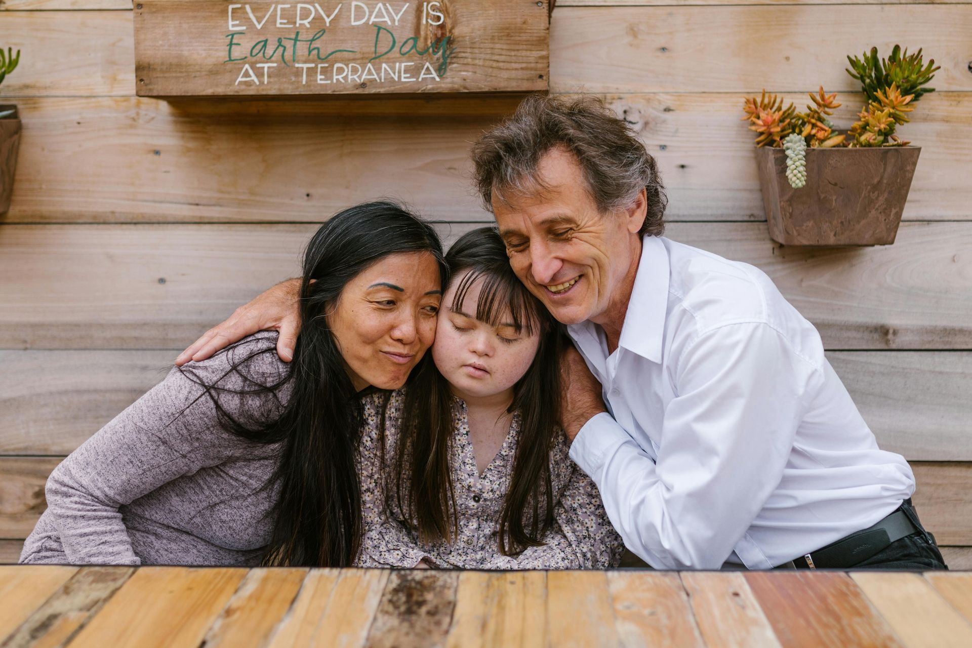 A family of three embrace closely against a wooden wall with potted plants, smiling warmly together.
