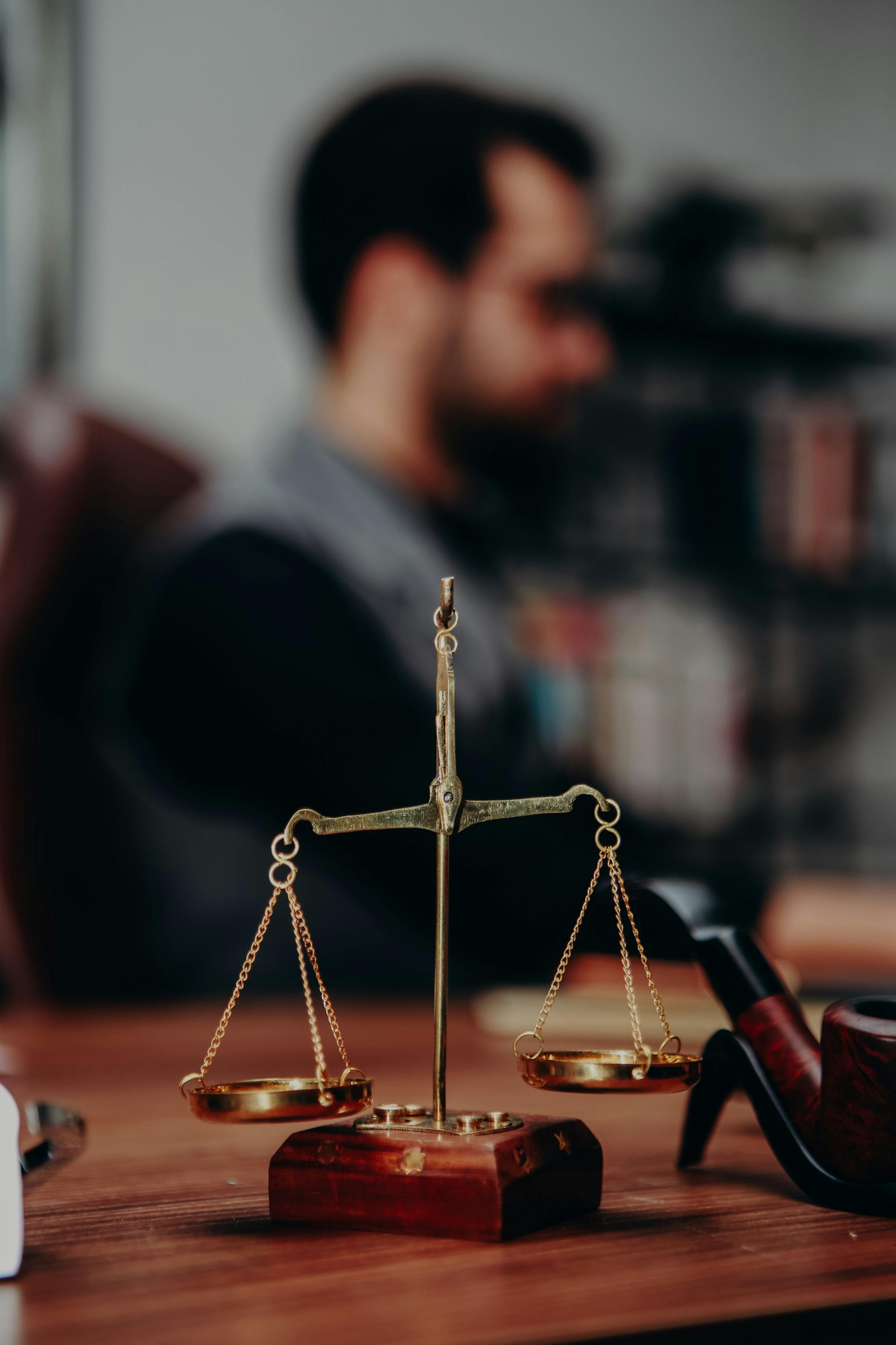 A gold-colored scale of justice sits on a wooden desk, with a blurred person in the background.