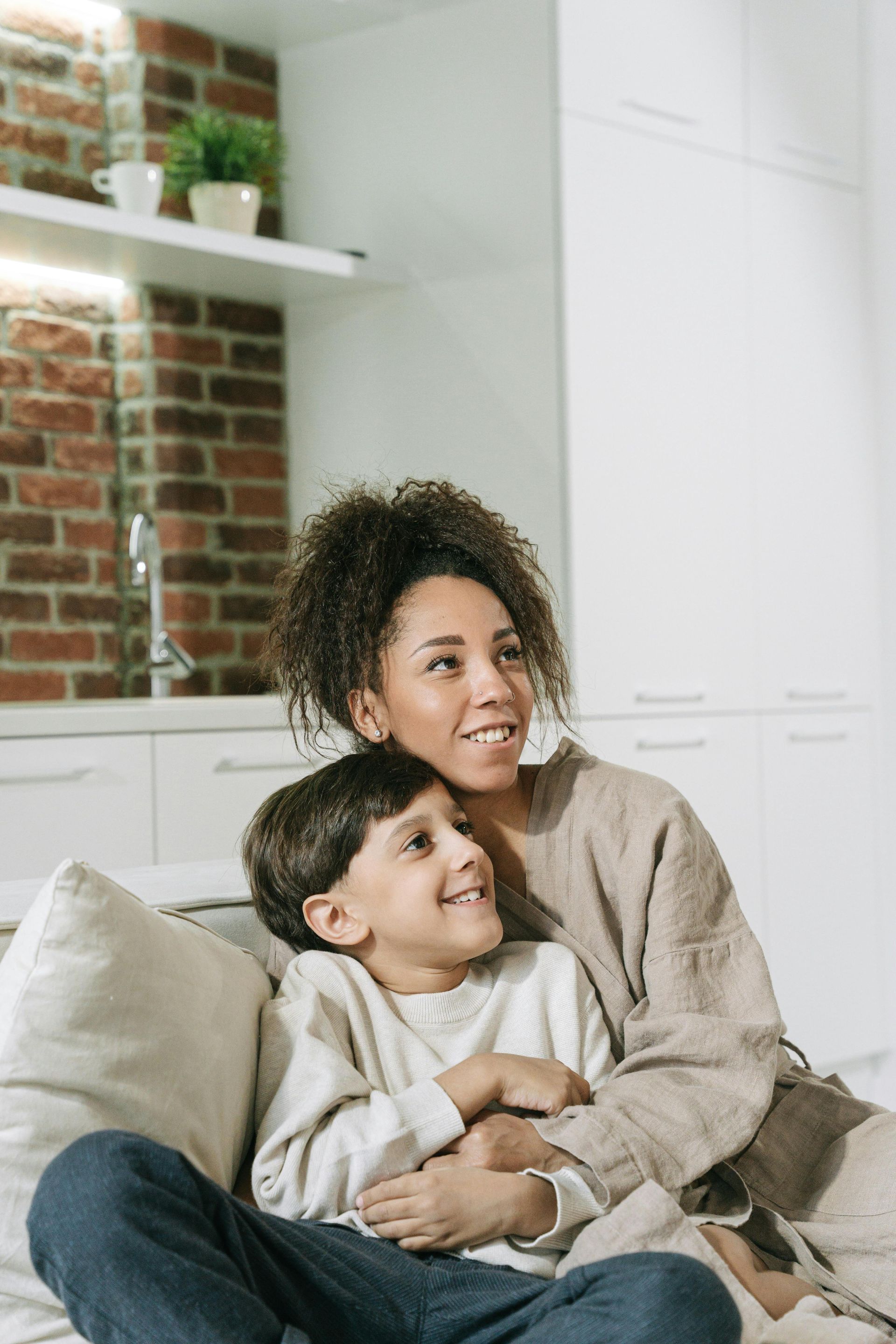 A woman and a child smiling together while sitting on a couch in a modern kitchen.
