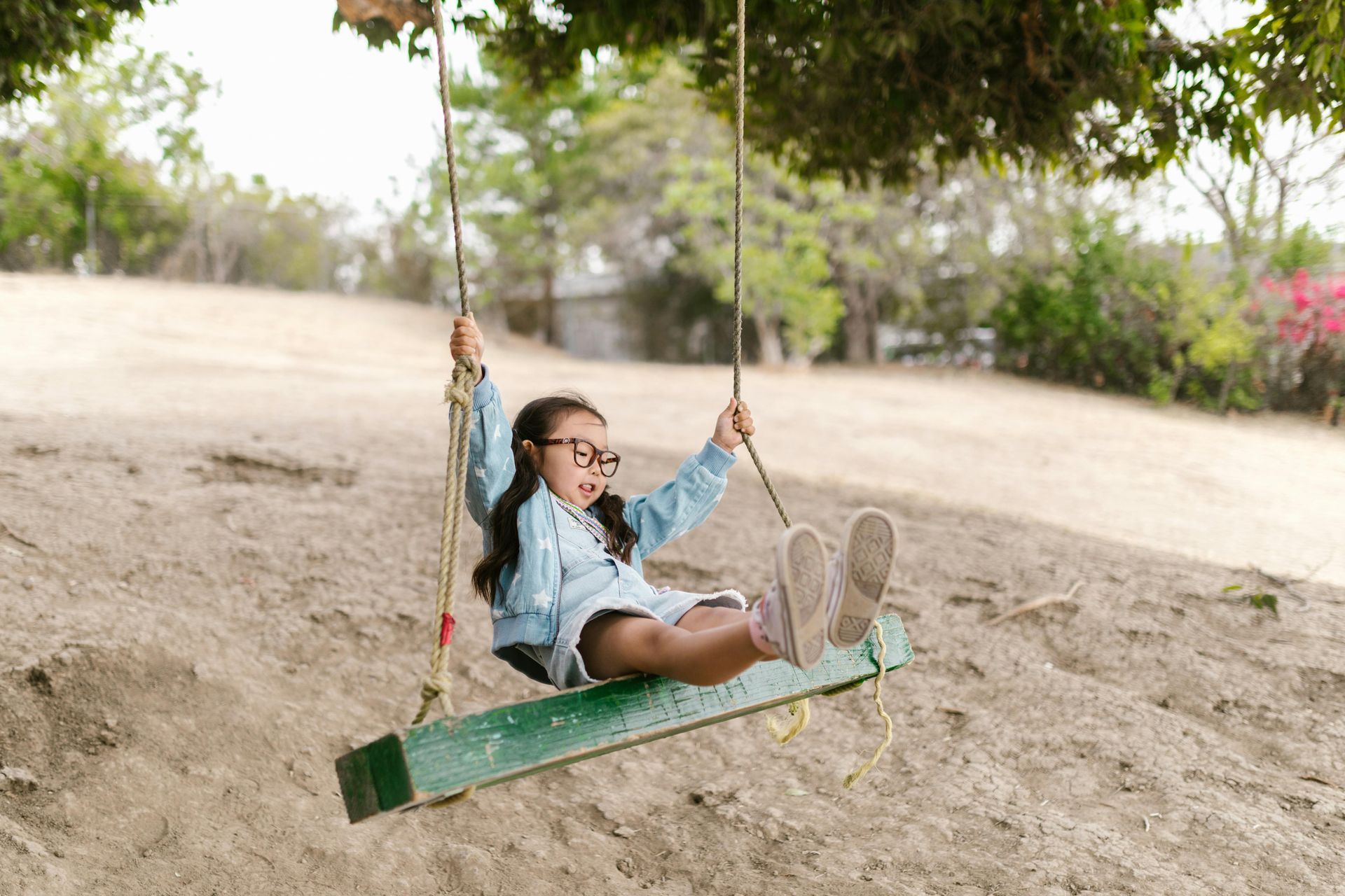 A child wearing glasses and a light denim jacket sits on a green wooden swing hanging from a tree in a sandy field.