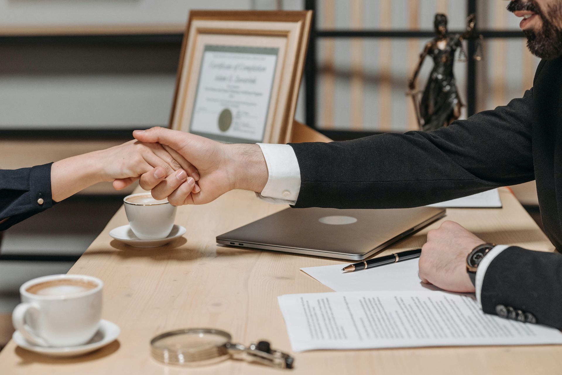 Two people shaking hands over a desk with coffee, a laptop, legal documents, and a statue of Lady Justice in an office.