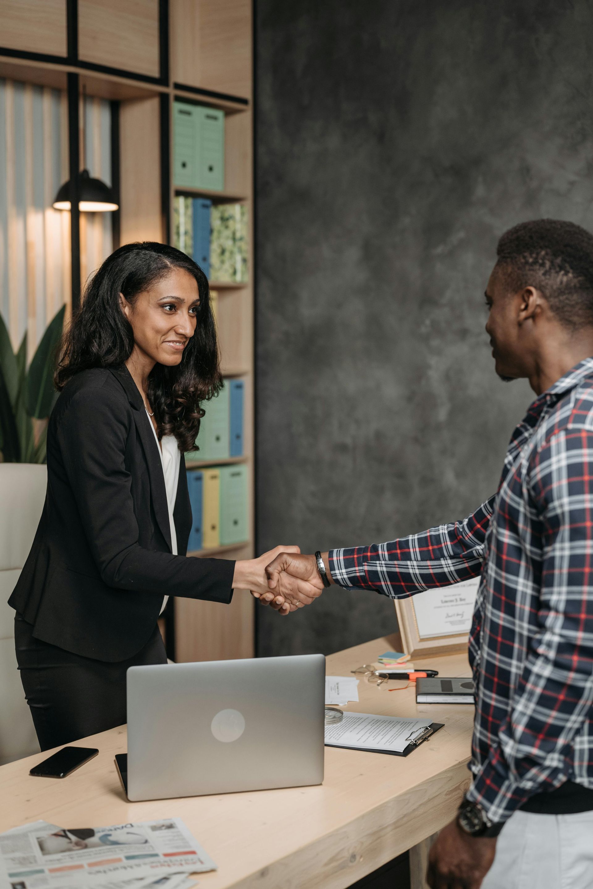 Two colleagues shake hands across a desk in an office setting.