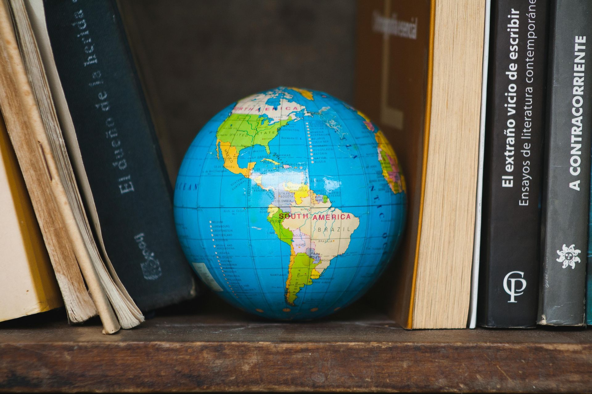 A small globe sitting on a wooden shelf between several hardcover books.