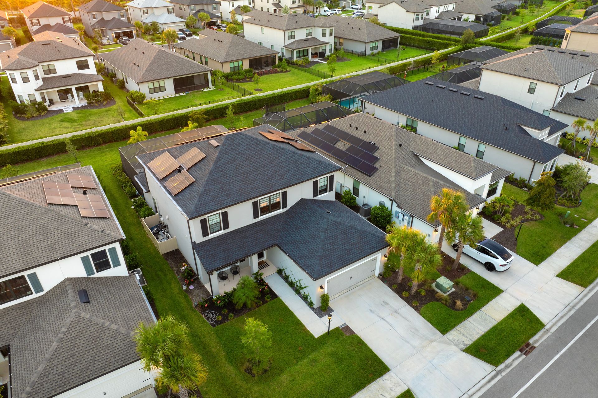 An aerial view of a suburban neighborhood with rows of modern houses featuring dark roofs, green lawns, and a white car.