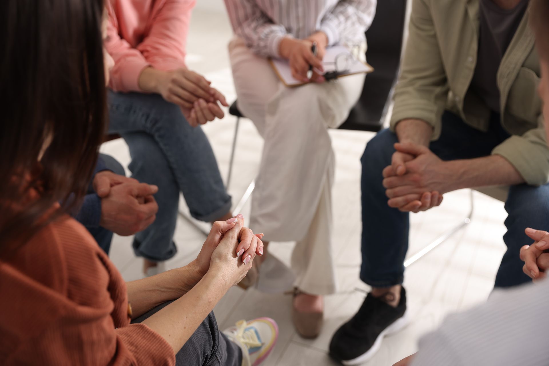 A diverse group sits in a circle, hands clasped, participating in a therapy or support group session.