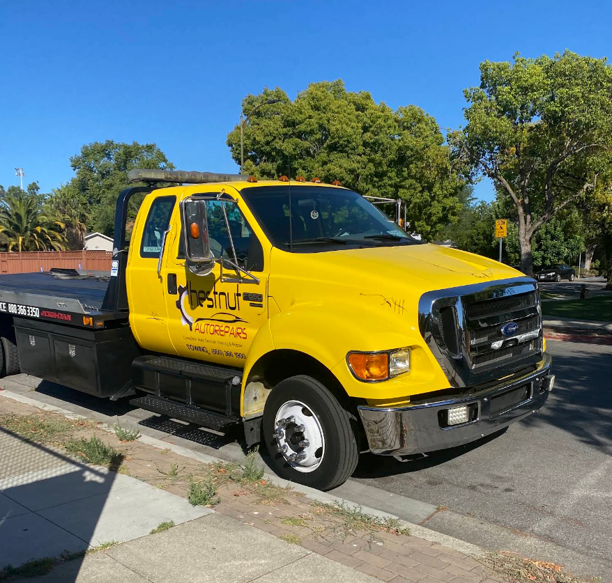 Yellow tow truck parked on a street beside a curb and trees, with blue sky in the background | Chestnut Auto Repairs & Towing