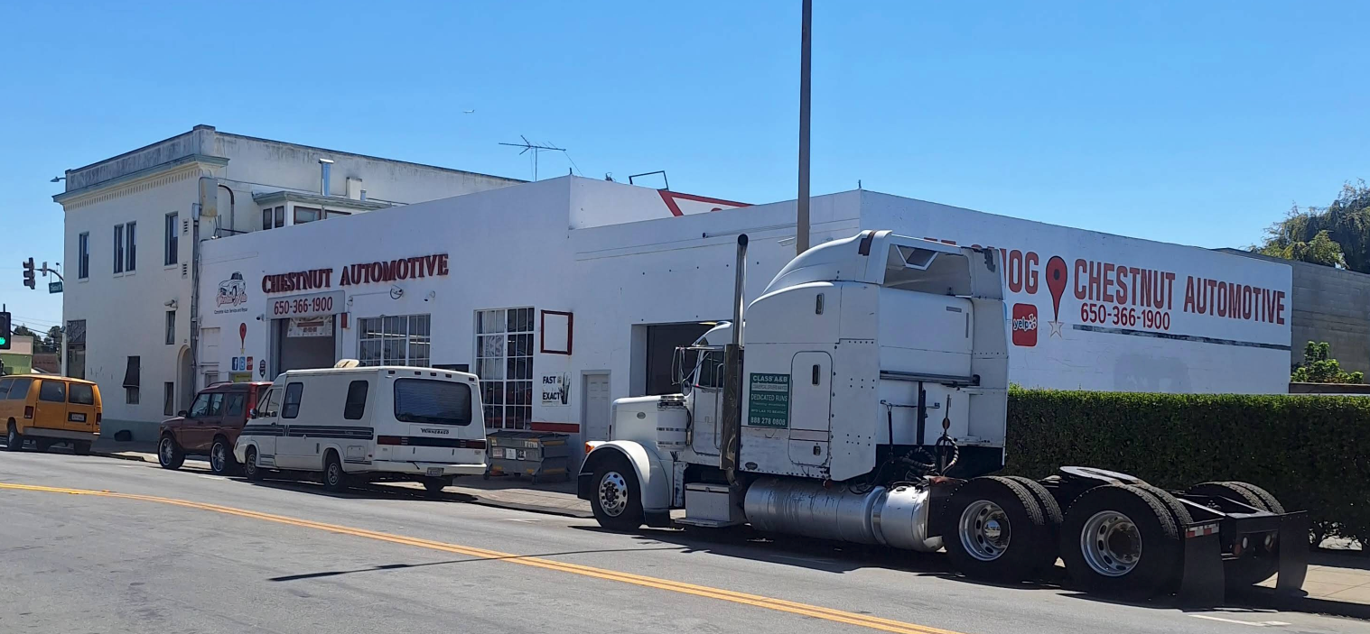White commercial building with parked vehicles and a semi-truck on a sunny street | Chestnut Auto Repairs & Towing