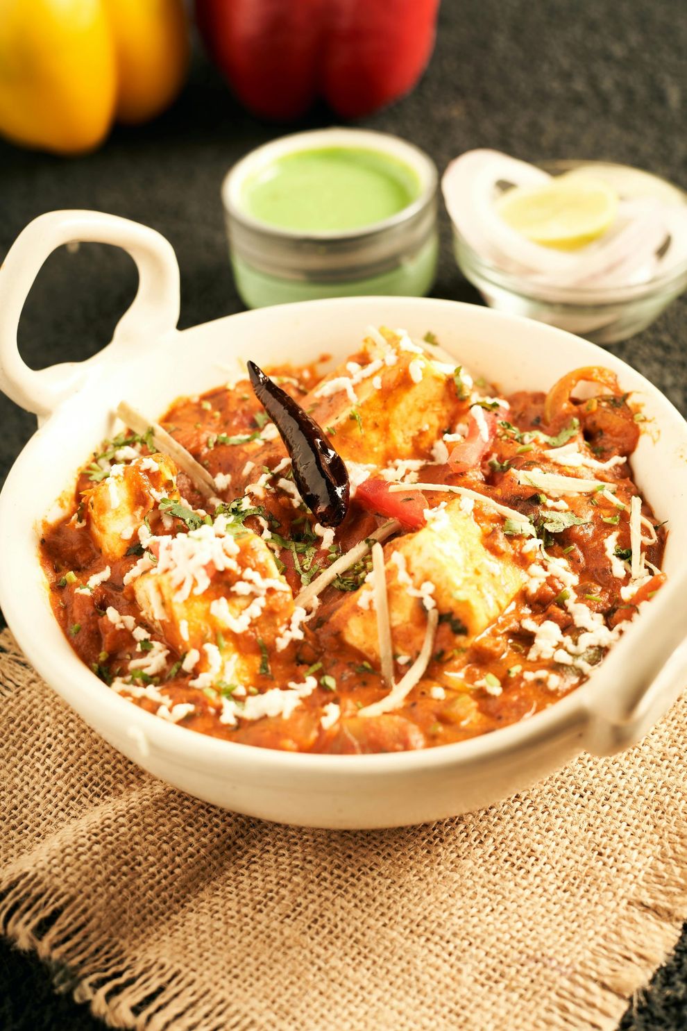 White bowl of red-orange curry topped with onions and herbs, on a burlap cloth with small condiment bowls nearby