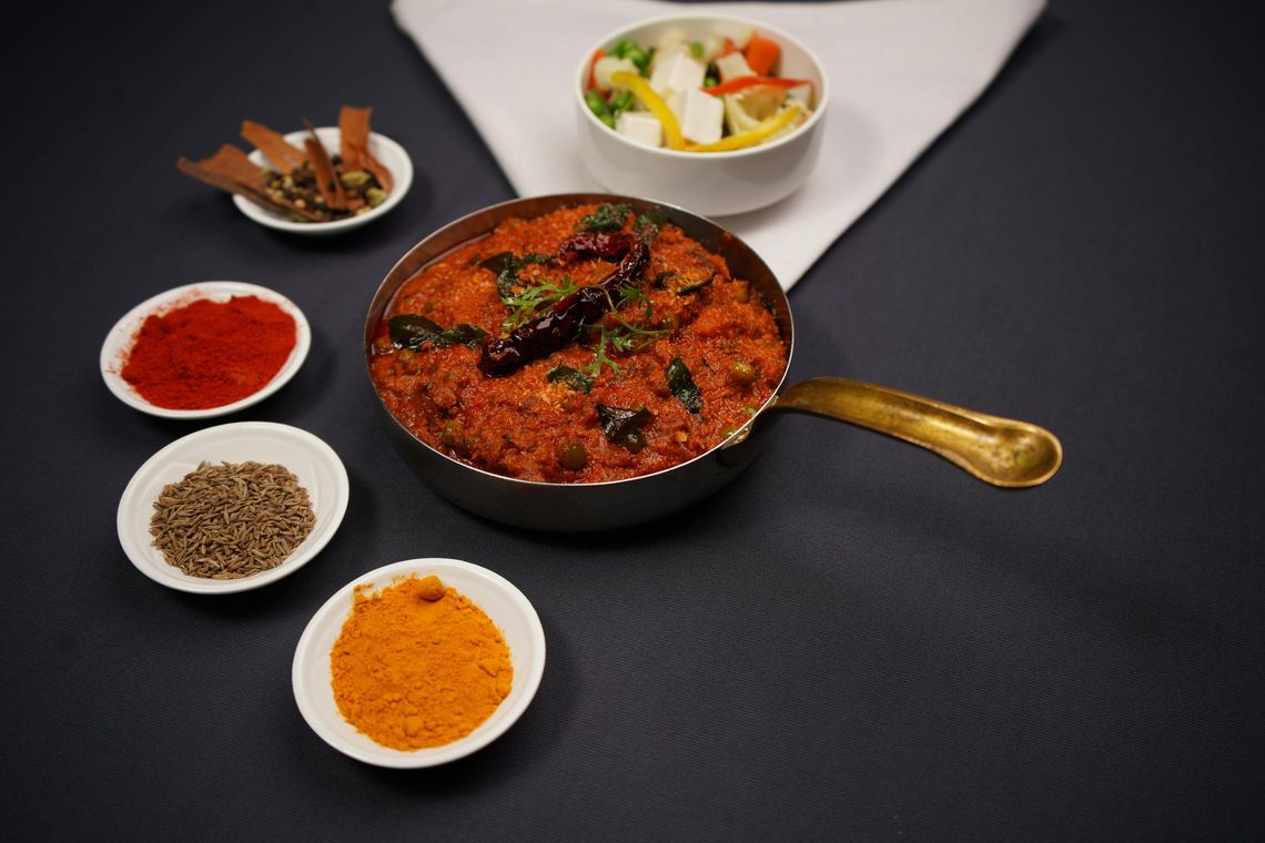 Spiced curry in a black pan with a brass handle, surrounded by small bowls of spices and vegetables