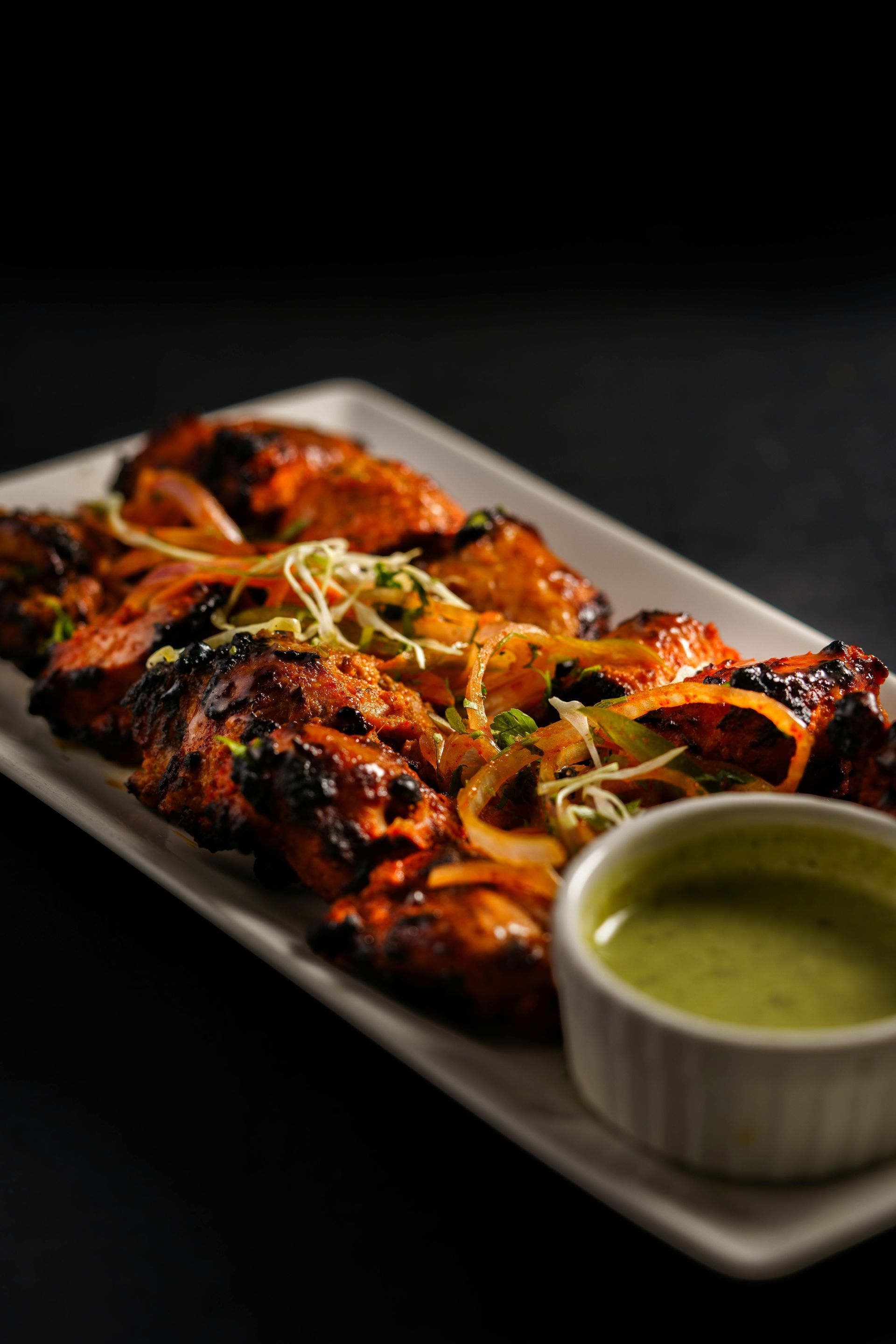 Grilled chicken skewers on a white platter with green dipping sauce, garnished with herbs.