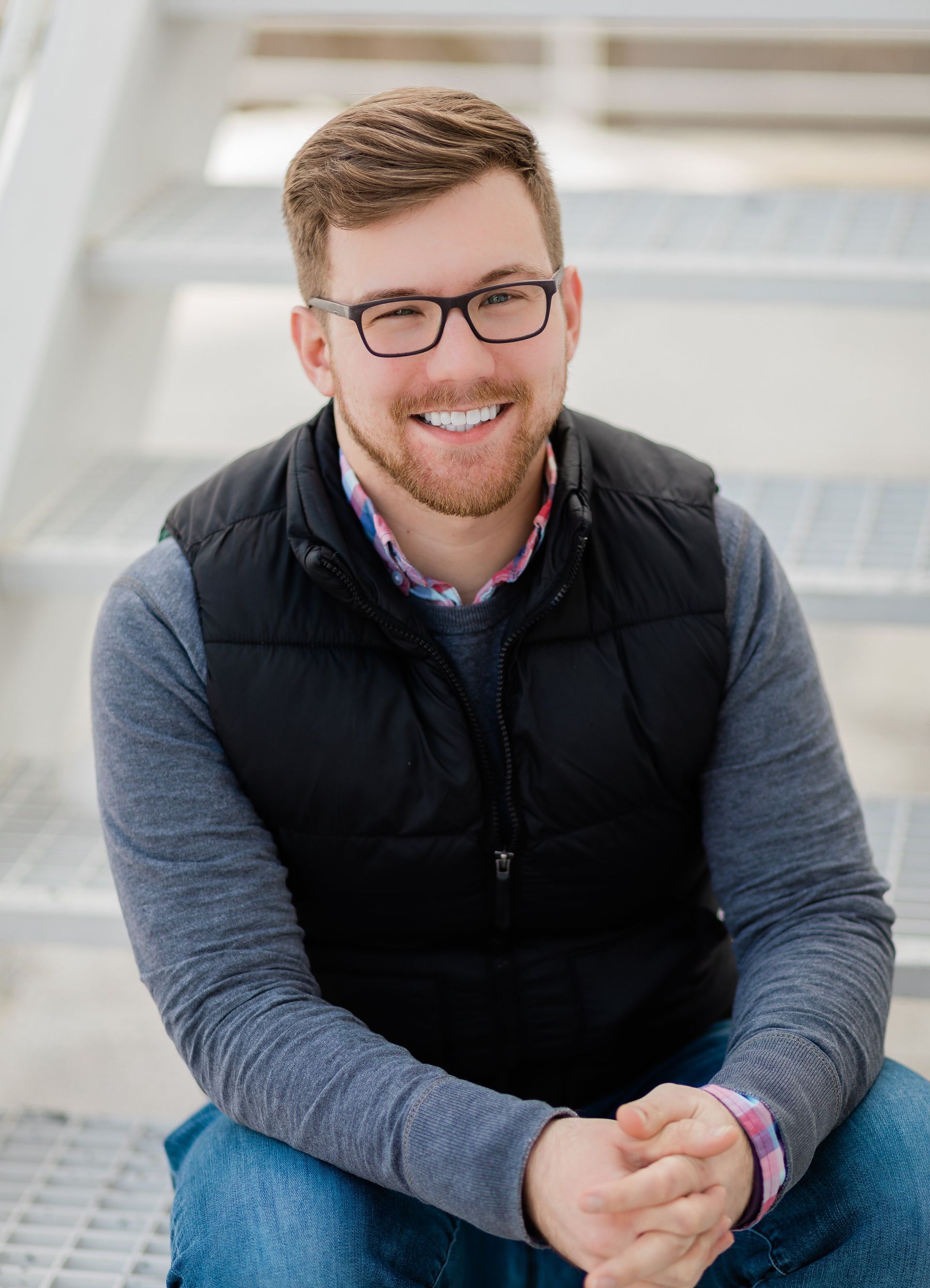 Man in glasses and vest smiles while sitting on metal stairs.