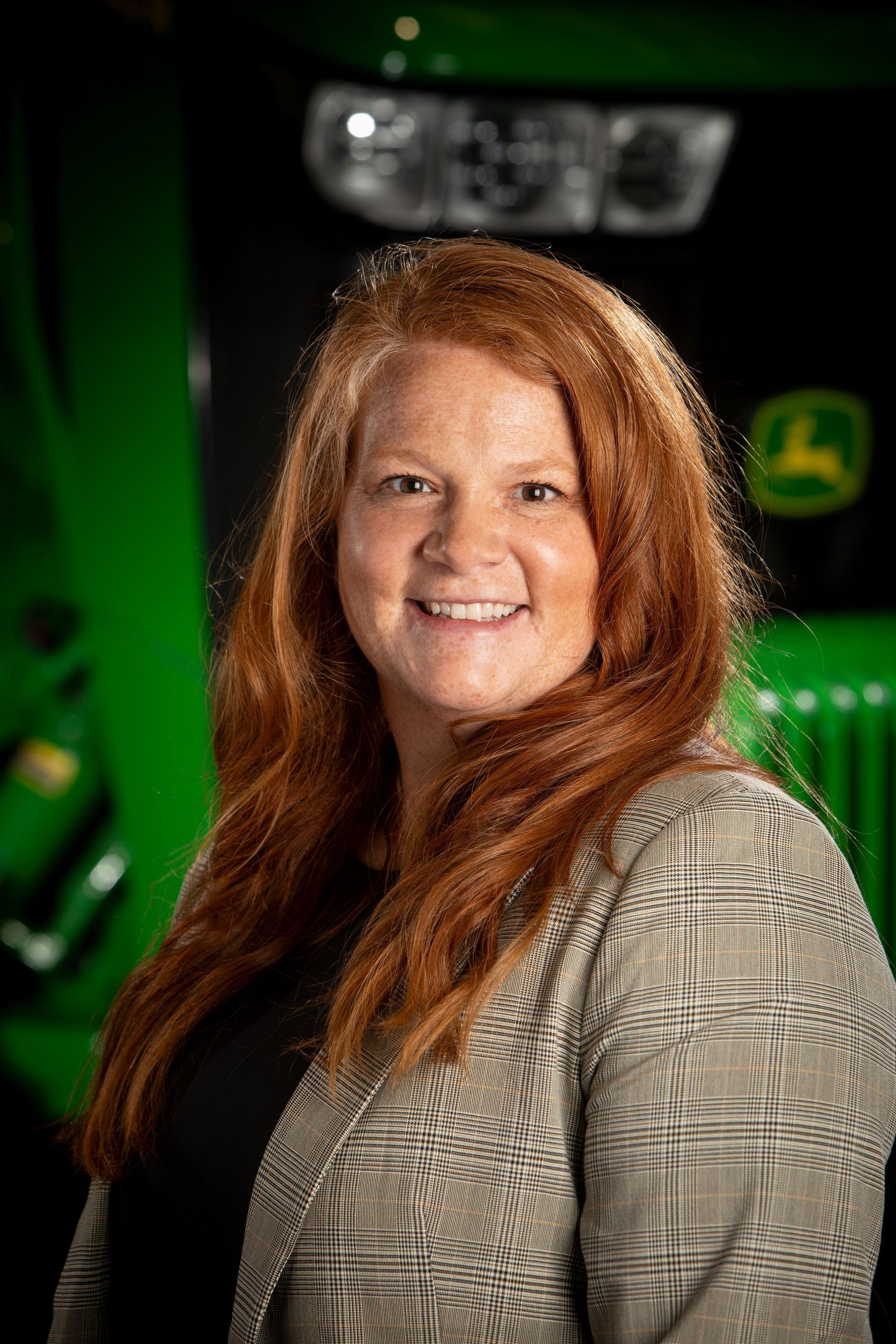 Woman with red hair smiling, wearing a blazer, standing in front of a green tractor.