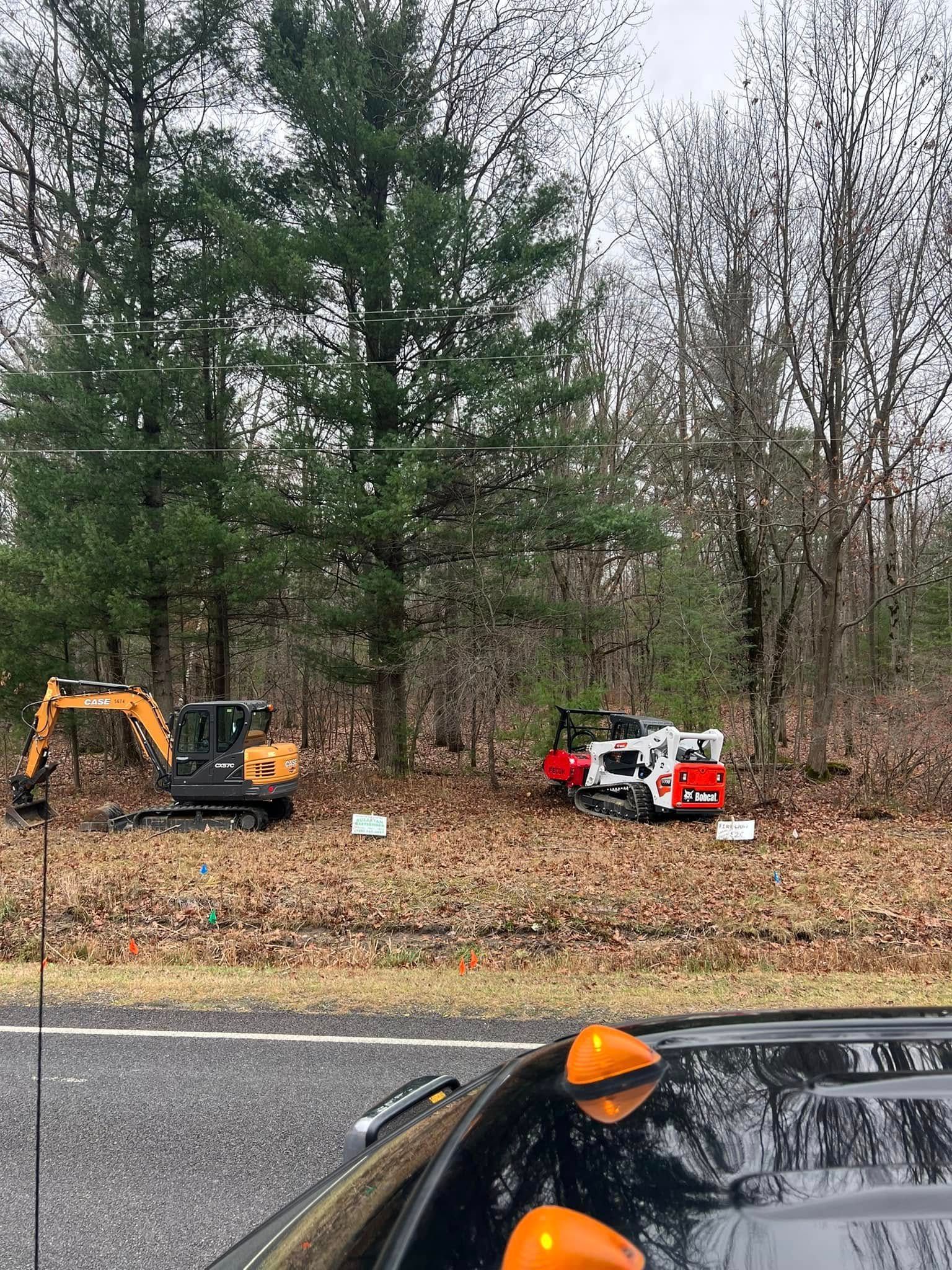 Construction equipment beside a road with a line of trees. An excavator and a white skid steer sit in a wooded area.