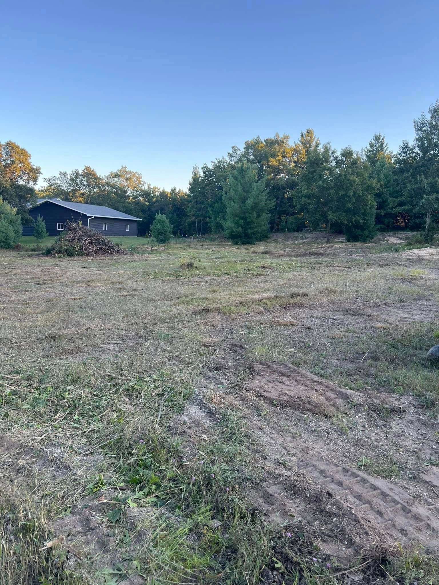 Cleared lot with sparse vegetation, small house in the background, surrounded by trees under a blue sky.