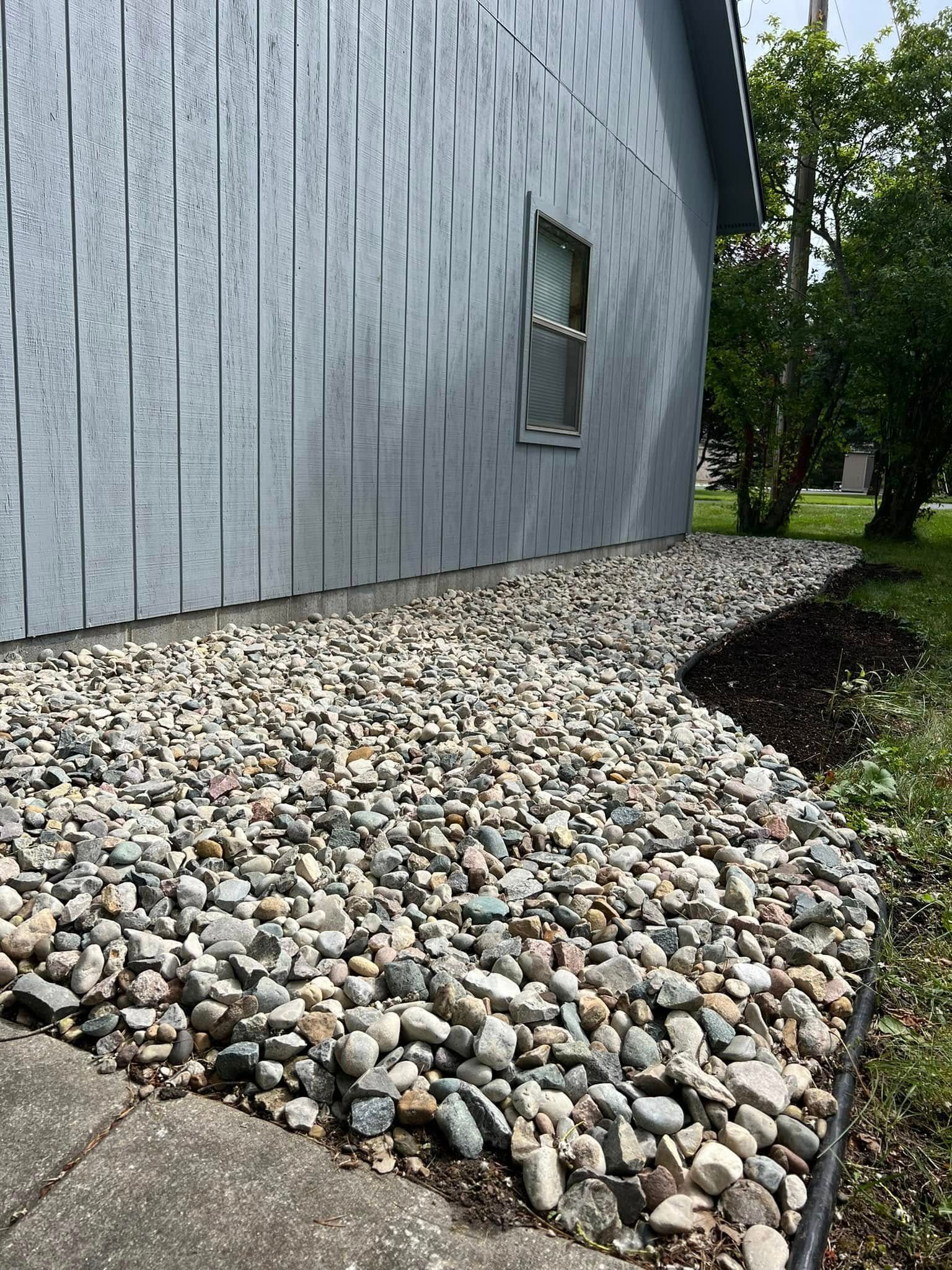 Gravel landscaping along a gray house exterior, next to a sidewalk and grass.