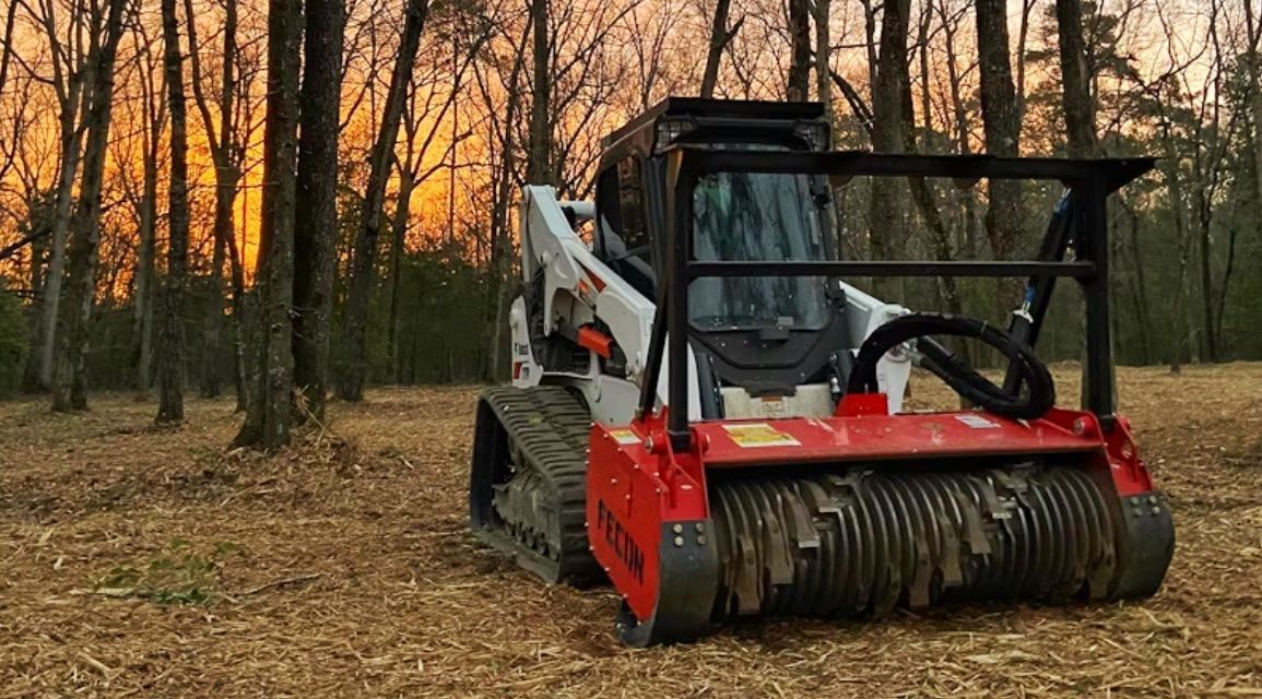 Skid steer with a forestry mulcher in a wooded area at sunset.
