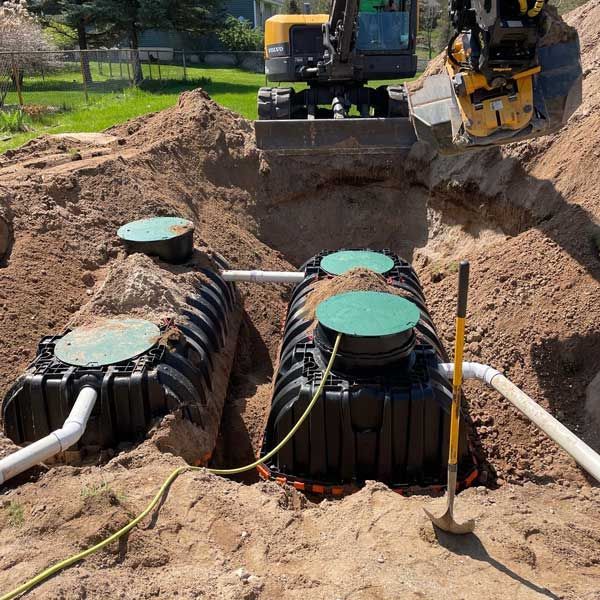Two black septic tanks in an excavated area, connected by pipes, with an excavator in the background.