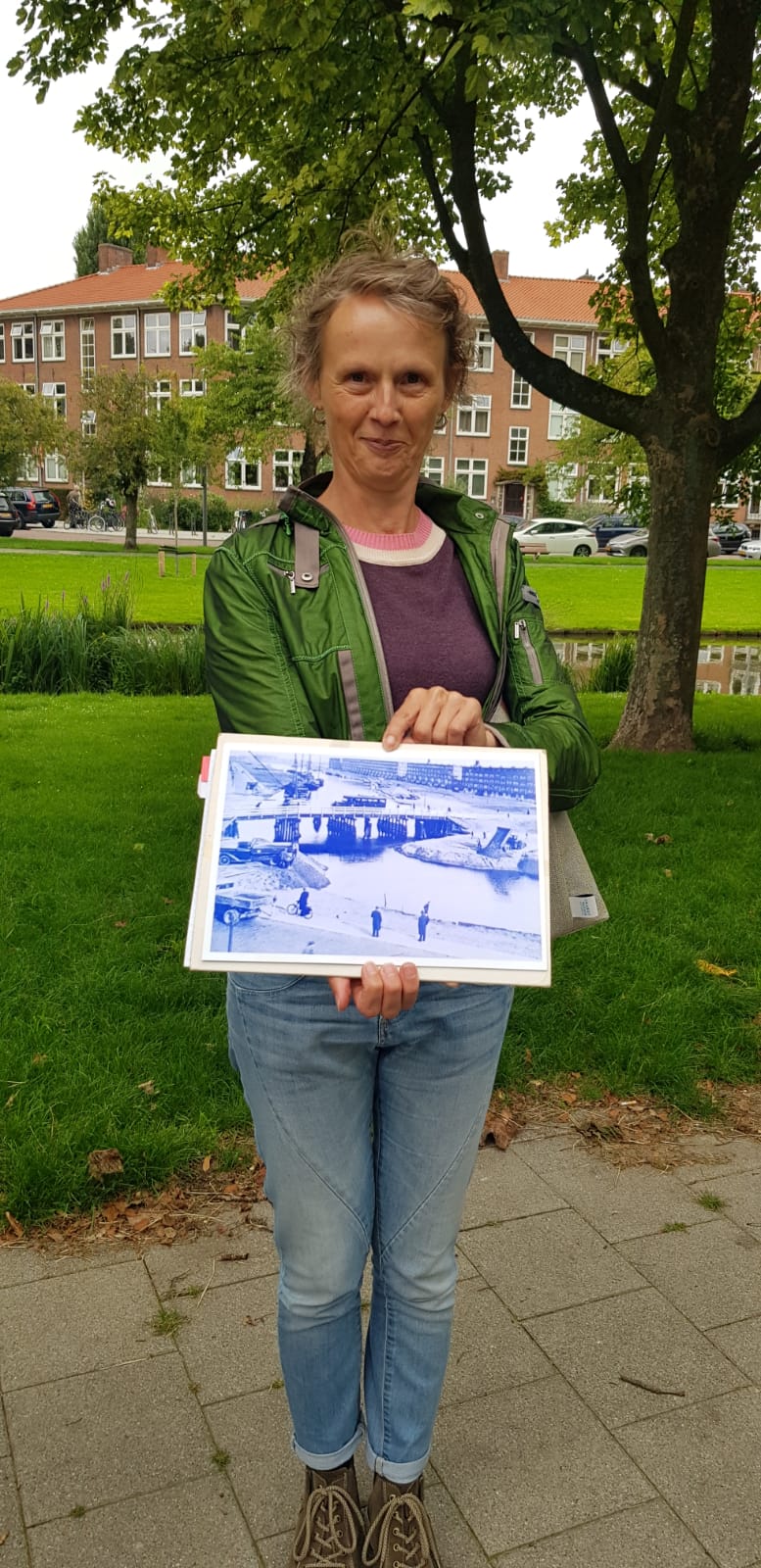 Vrouw in groene jas en spijkerbroek houdt buiten bouwtekeningen vast, met bomen en gebouwen op de achtergrond.