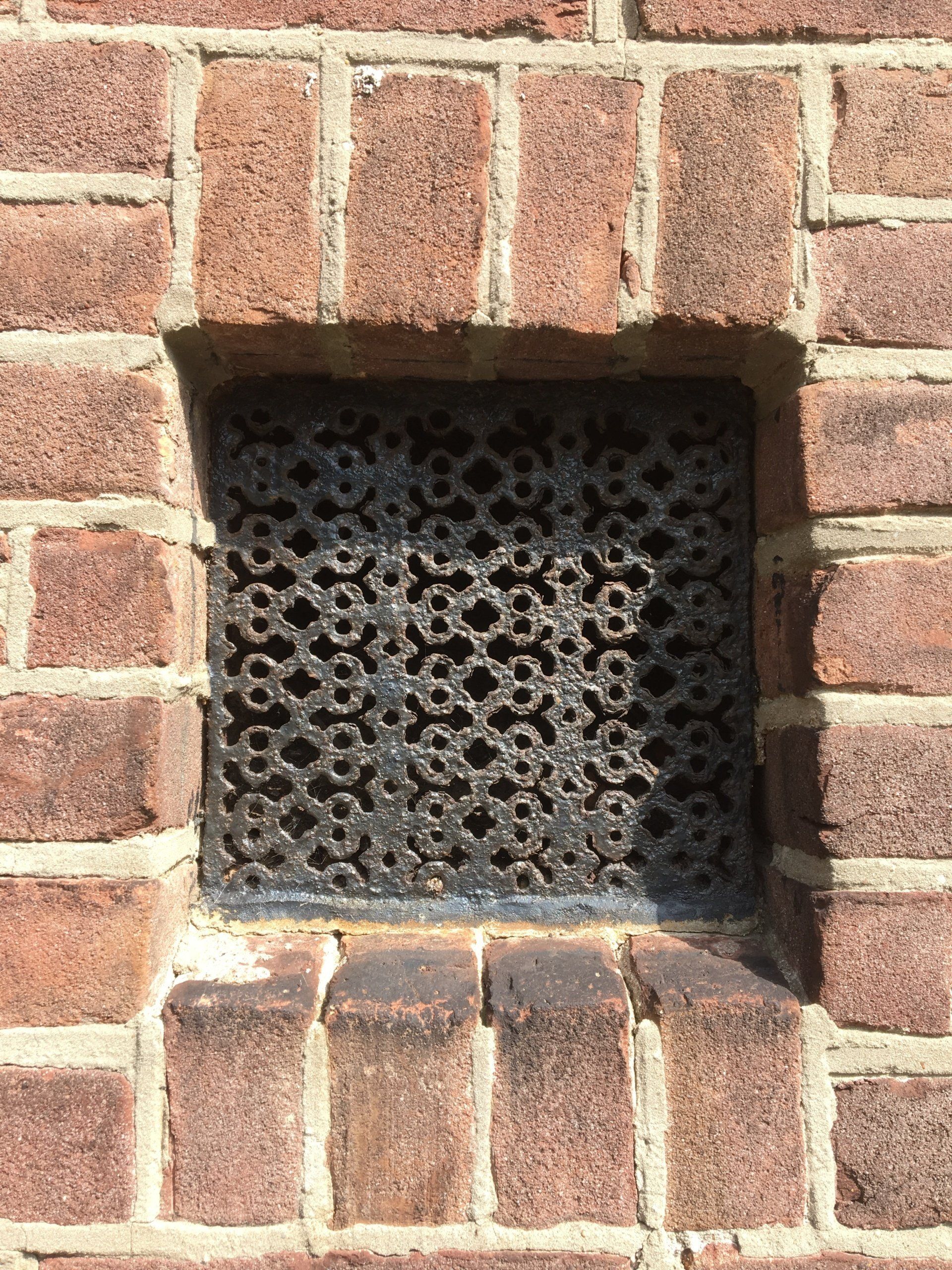 Bakstenen muur met een klein, decoratief metalen ventilatierooster.