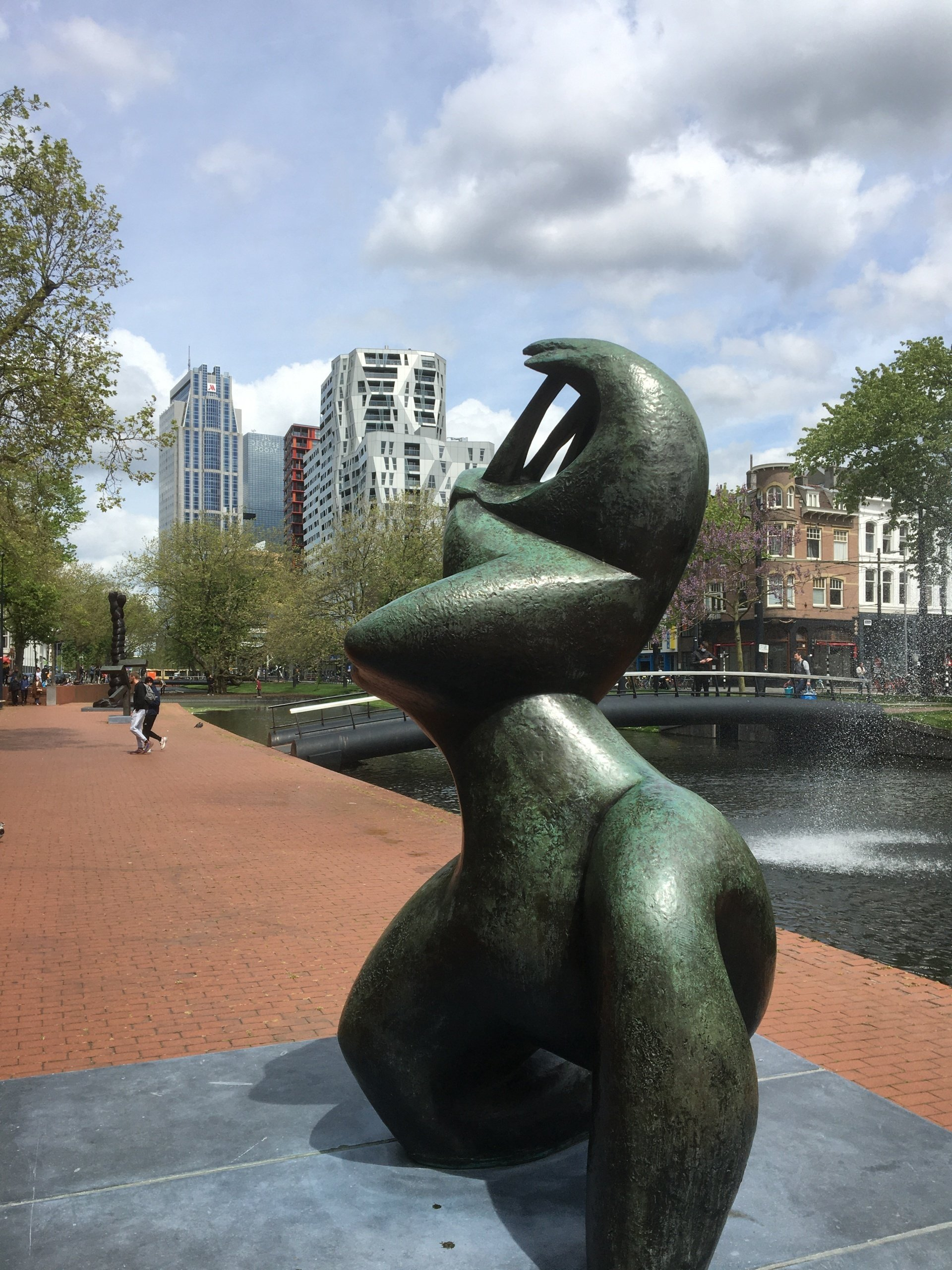 Bronzen sculptuur in een park, met moderne gebouwen en een kanaal op de achtergrond.