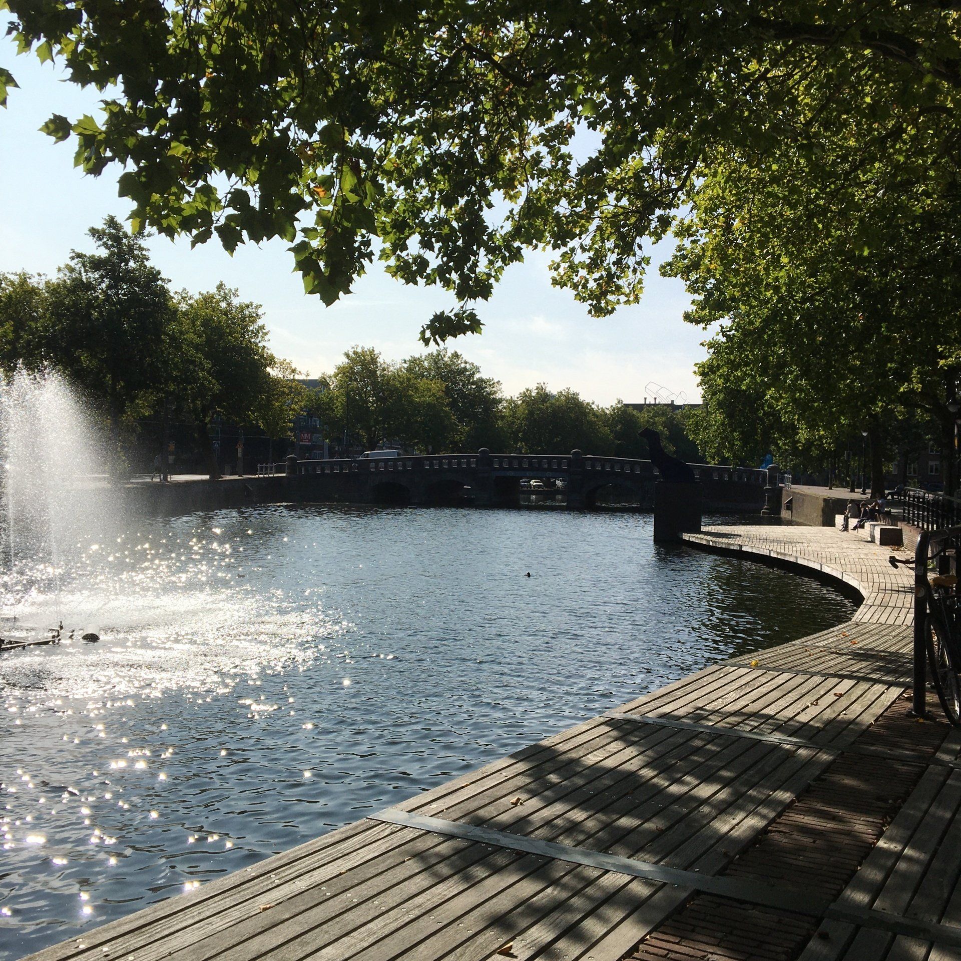 Een fontein spuit water in een meer. Zonlicht schittert op het water. Bomen omlijsten het uitzicht.