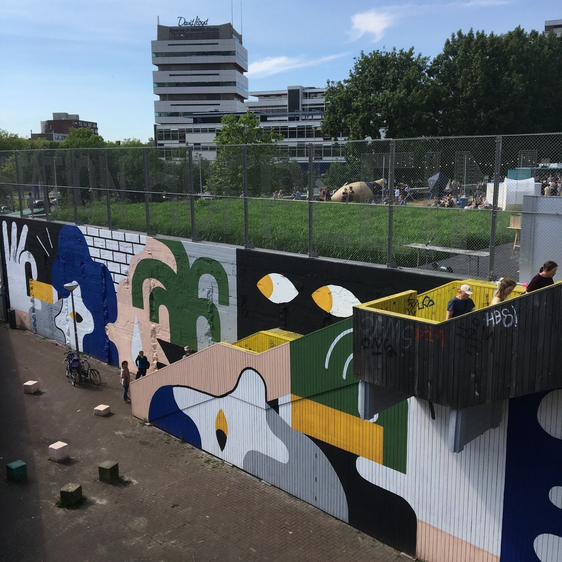 Kleurrijke muurschildering op een keermuur; park erboven. Mensen in het gras. Hoog gebouw op de achtergrond.