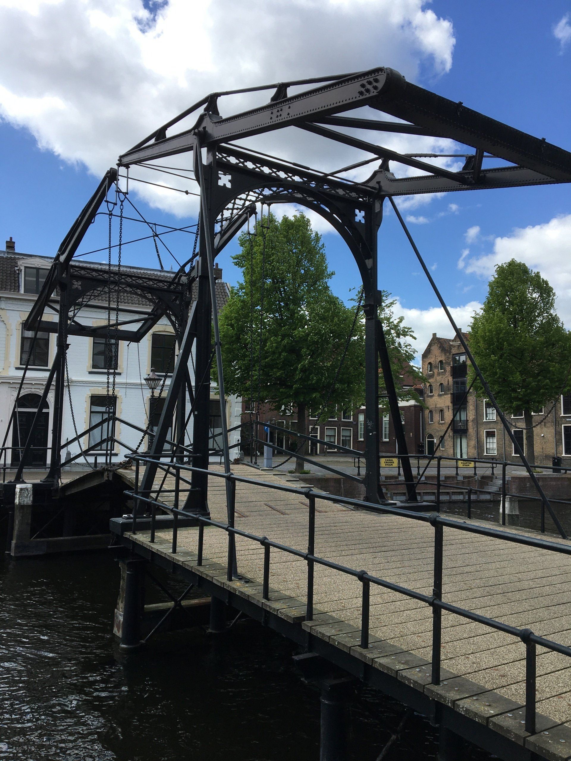 Een zwarte ophaalbrug over het water met een grijs pad en gebouwen op de achtergrond. Blauwe lucht met wolken.