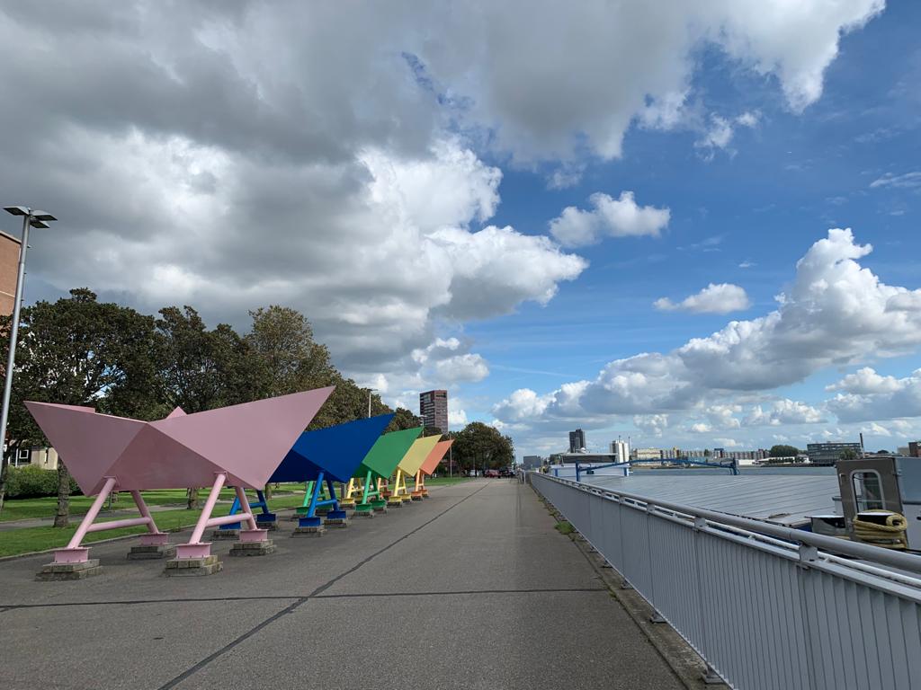 Kleurrijke origami bootsculpturen staan ​​langs een pad met op de achtergrond een stadsgezicht onder een bewolkte hemel.