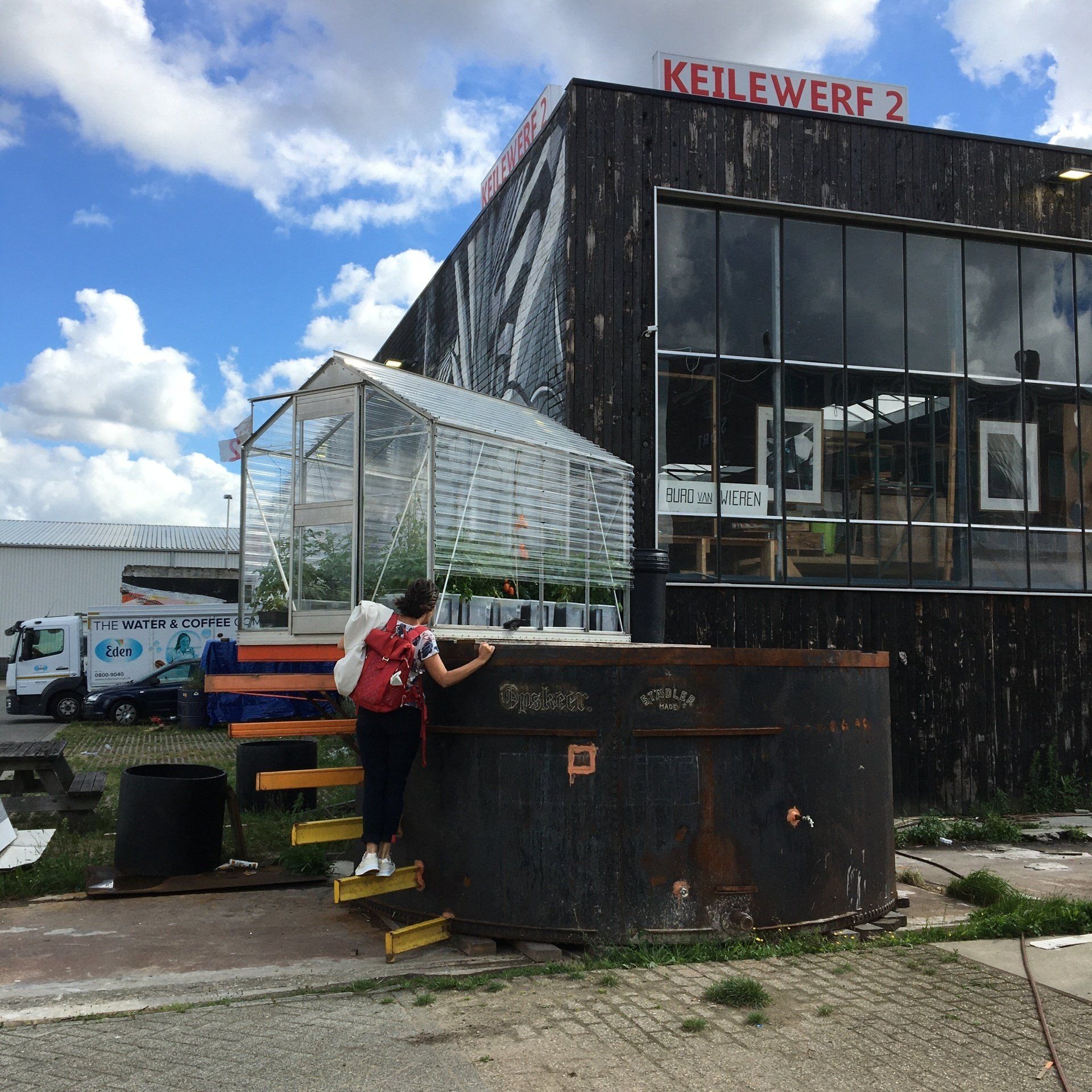 Een persoon beklimt de trap naar een kas. Op de achtergrond zijn een modern gebouw en een bewolkte hemel te zien.