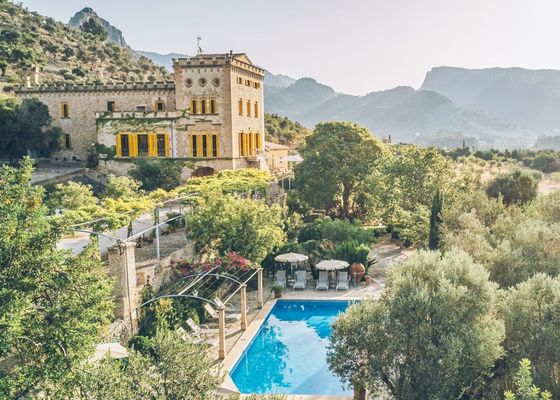 Una vista aérea de un castillo con una gran piscina frente a él rodeado de árboles y montañas.