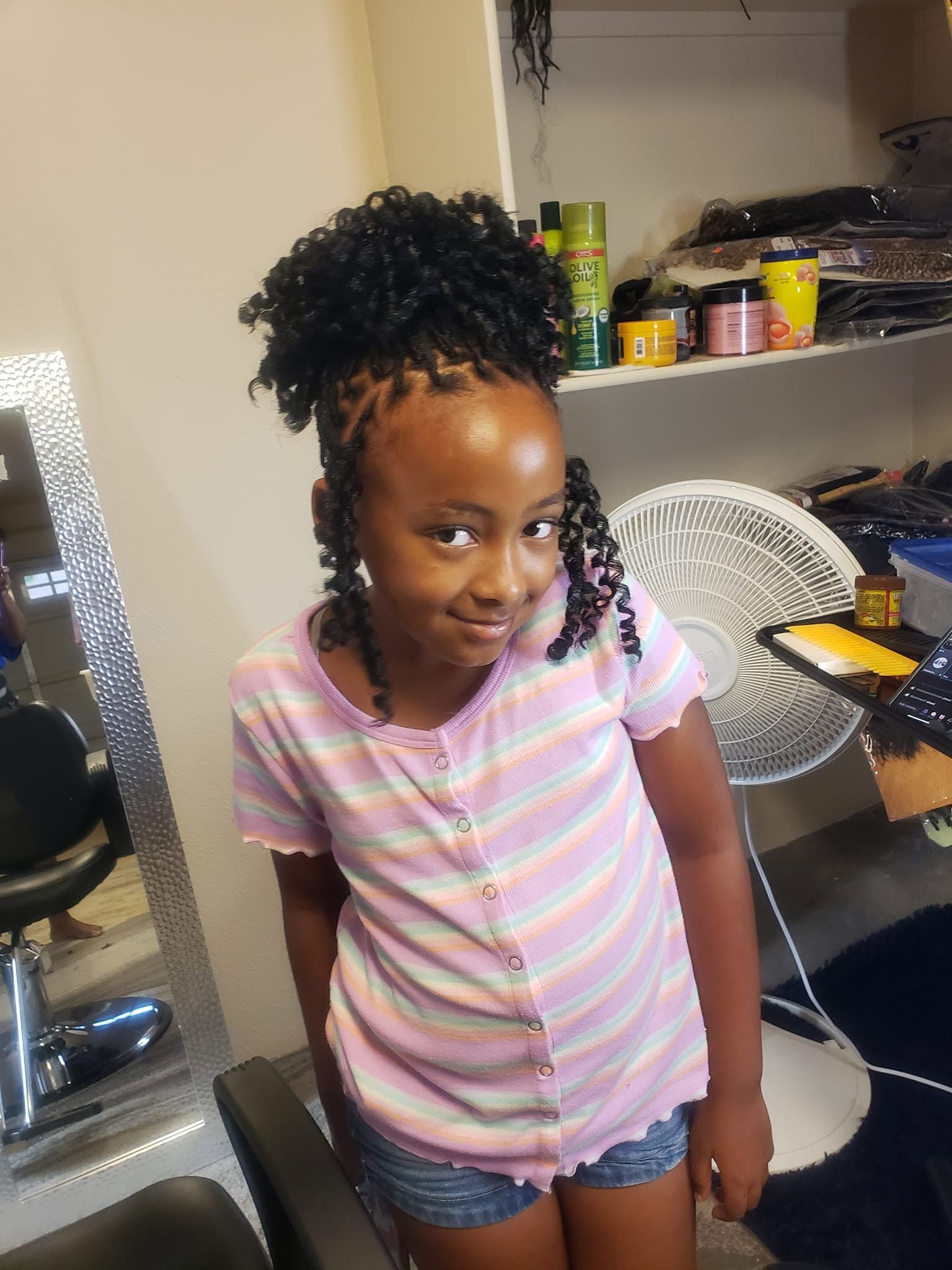 Child smiling in a pink striped shirt and denim shorts, standing in a room with a fan and shelves.