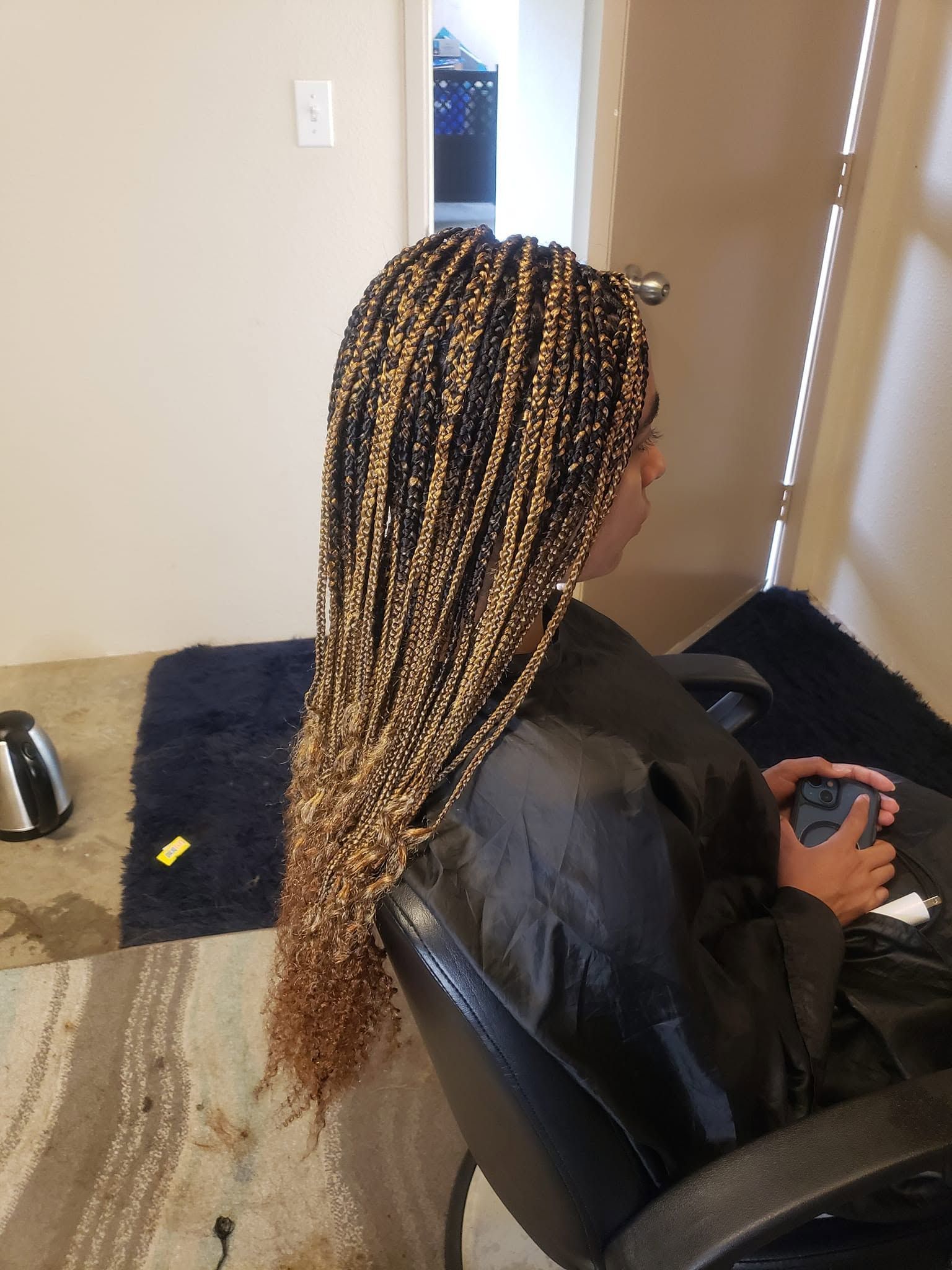 Person with long braided hair seated in a black chair, holding a phone in a room by a window.